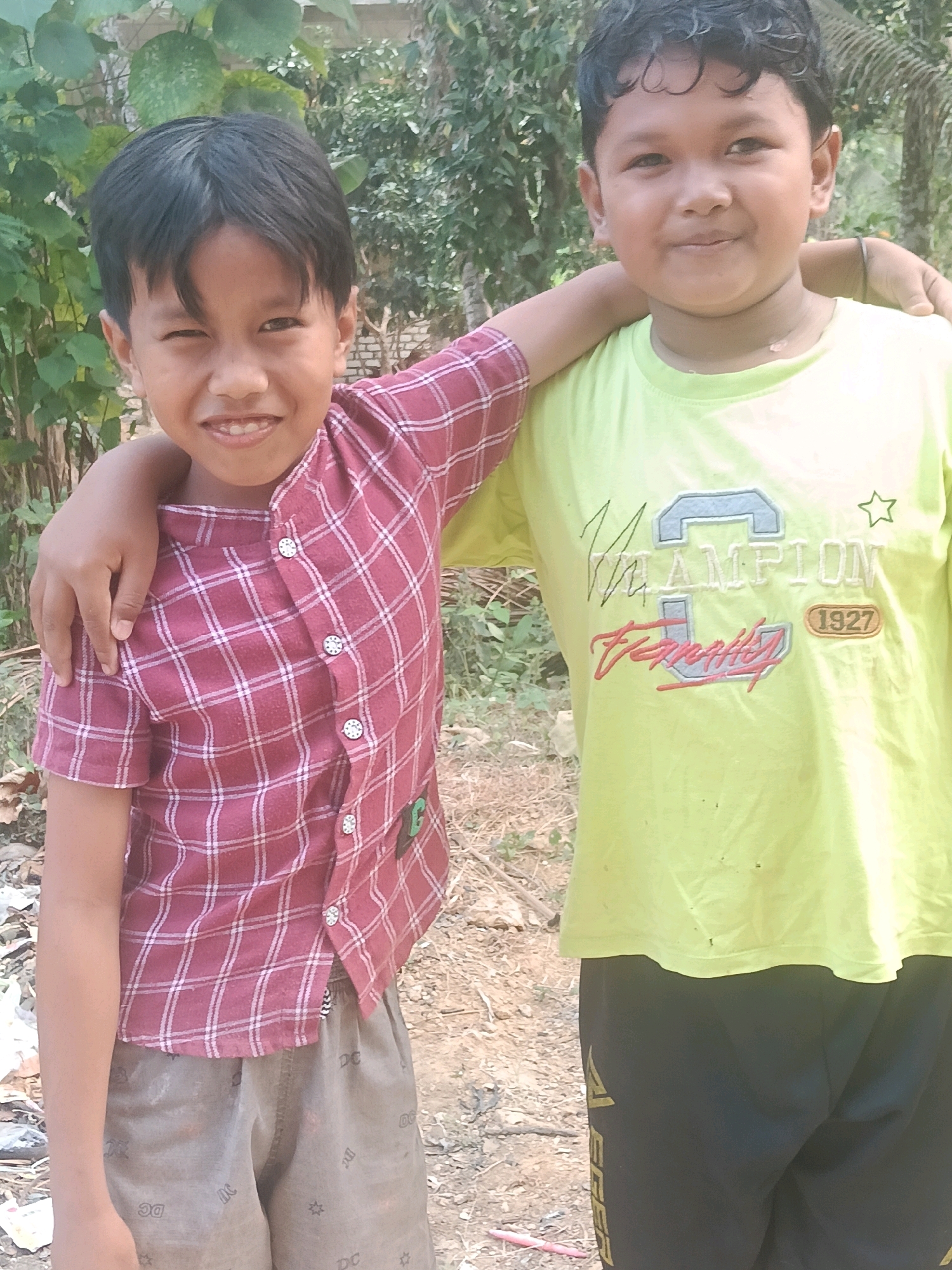 Two young boys, appearing to be of Southeast Asian descent, are standing close together outdoors. The boy on the left is wearing a red and white plaid shirt and brown shorts. He is winking and has his left arm around the shoulder of the other boy. The boy on the right is wearing a lime green t-shirt with "CHAMPION" and "1927" graphics, and black pants. He is looking at the camera with a slight smile. The background consists of lush green foliage and a dirt path, suggesting a rural or semi-rural setting. The scene is well-lit by natural light, indicating it was taken during the daytime. There are no visible buildings or distinct urban features that would specifically place the image in Bulangan Barat, Indonesia.