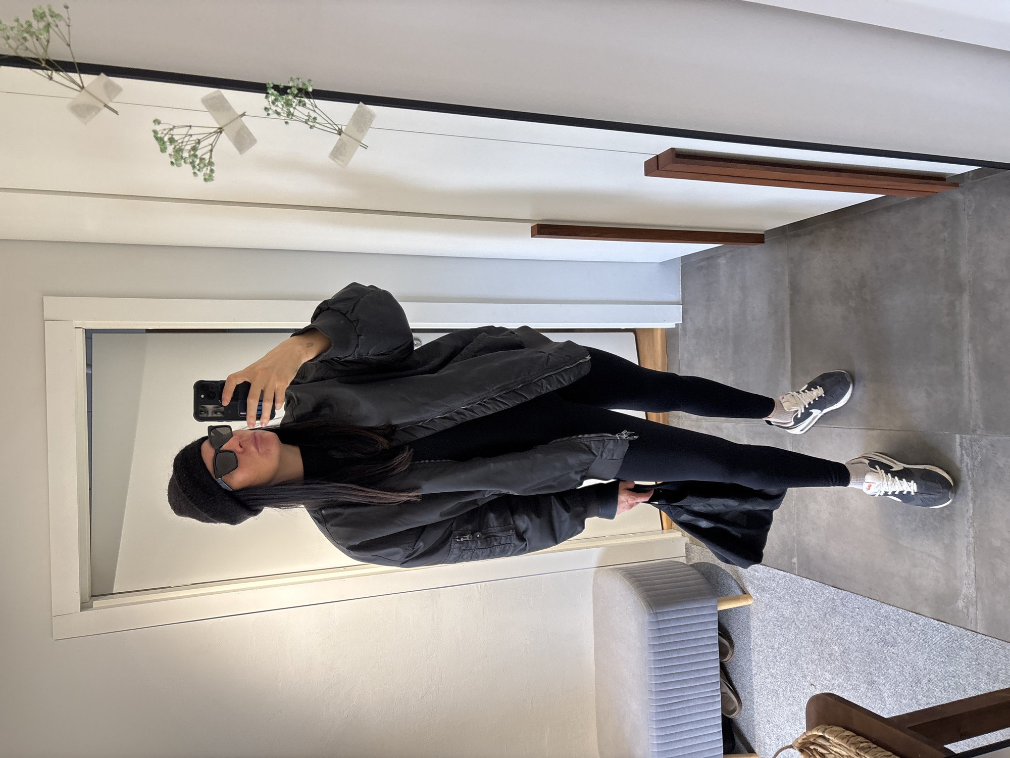 A person is captured in a mirror selfie within what appears to be a modern indoor setting, likely a hallway or entryway in Chaikovsky, Russia. The individual is dressed in an all-black, casual outfit, consisting of a black beanie, black sunglasses, a loose-fitting black bomber jacket, and black leggings. They are wearing dark grey sneakers with white soles and laces, paired with white socks. The person holds a black smartphone with a black case up to take the photo, partially obscuring their face.
The reflected background features a minimalist interior design. The floor is covered with large, light grey rectangular tiles, and the walls are white. To the left, a white door frame is visible. Decorative elements above the person include three small sprigs of baby's breath flowers, which are taped to a thin string or wire stretched across the upper part of the mirror. Further right, two horizontal wooden beams or slats are affixed to the wall or ceiling. In the lower right foreground of the reflection, a light grey upholstered bench with a ribbed texture and wooden legs is partially visible, next to a wooden stool or table topped with a woven basket. The overall impression is one of modern, clean aesthetics. There is no visible text in the image.