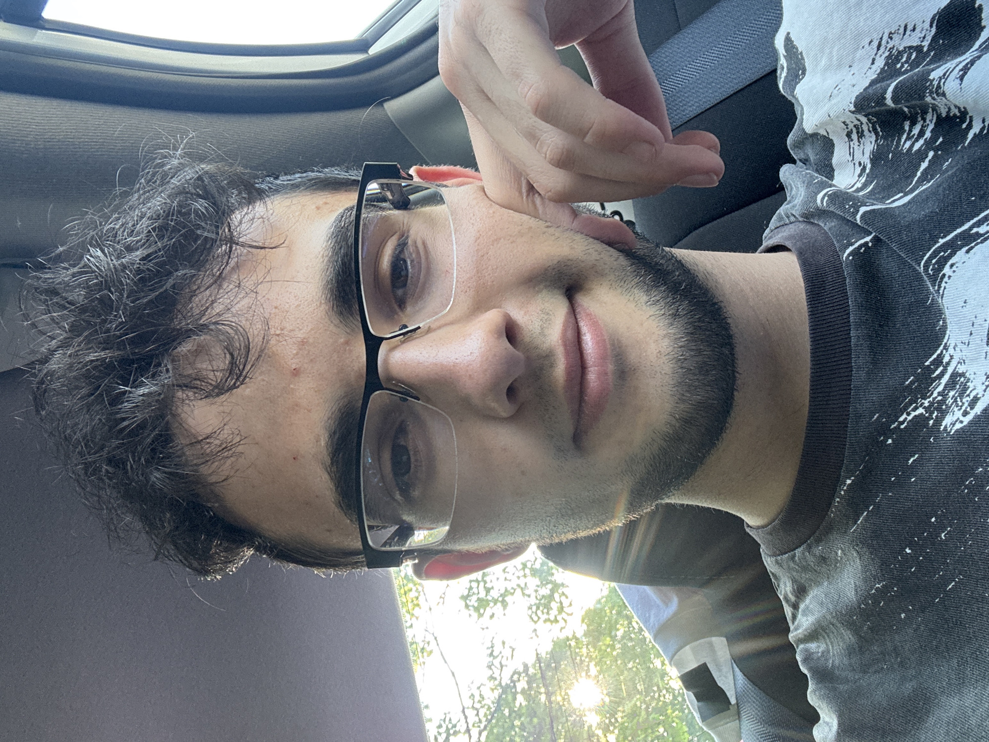 A close-up shot of a man with dark hair, glasses, and a beard. He is wearing a patterned shirt and appears to be inside a vehicle. The sun is visible through the windshield. The location may be Franklin Township, USA, given the prompt's context.