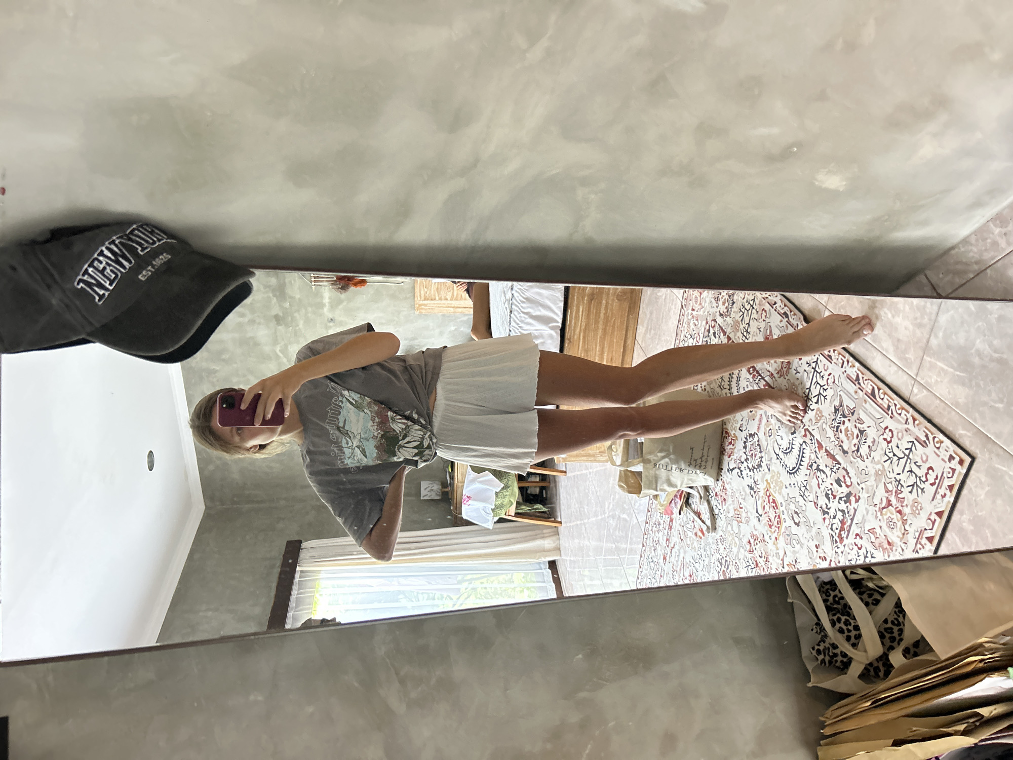 A woman takes a selfie in a mirror, likely in a home in North Kuta, Indonesia. The woman is wearing a grey t-shirt and white shorts. A grey baseball cap hangs on the mirror. In the background, a rug with a pattern, a window, a bed, and a few bags are visible. The environment appears to be a domestic space, perhaps a bedroom or living area.
