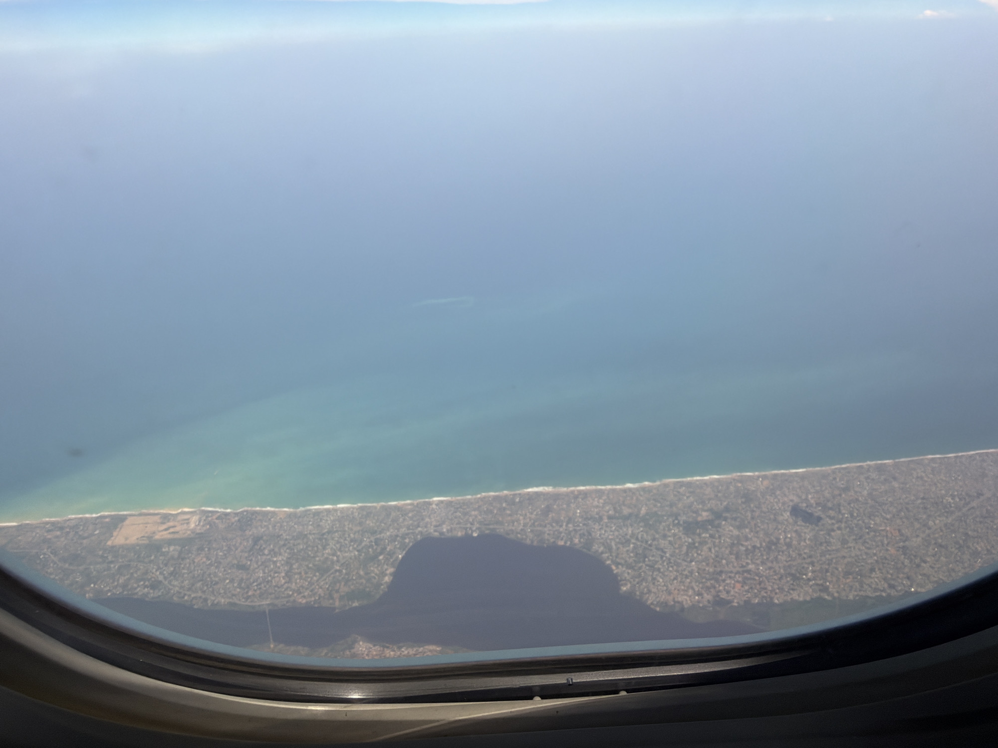 The image shows a view from an airplane window overlooking a coastal area. The ocean fills the upper portion of the frame, transitioning to a shoreline with a dense urban environment along the coast. The city is likely Cotonou, Benin, based on geographical cues and the nature of the urban layout visible.
Specifically, the shoreline exhibits a narrow sandy beach followed by densely packed buildings and infrastructure stretching inland. There is a prominent dark area in the lower central portion of the window view, likely caused by a shadow or reflection within the window itself. The water color shifts from a deeper blue further out to a lighter turquoise near the shore, indicating shallow water. The sky is bright with some hazy conditions near the horizon.