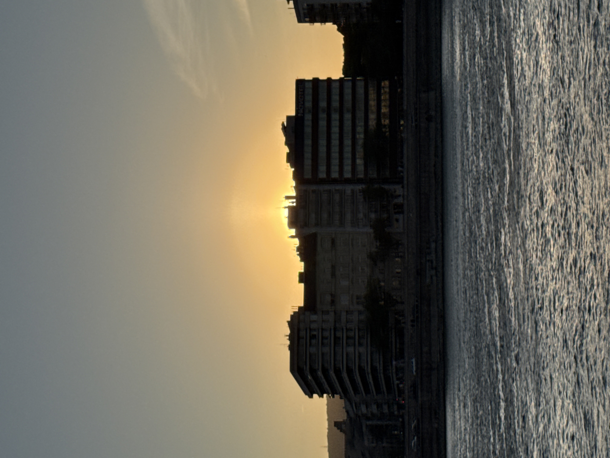 The image depicts a striking urban waterscape at either sunrise or sunset, strongly suggesting the Danube River in Budapest, Hungary. A dramatic skyline of buildings stands silhouetted on the left against a brilliant sky, which transitions from an intense, warm orange and yellow near the horizon to a hazy blue-grey higher up. The sun, a powerful source of light, is positioned low behind some of the structures, creating a stark contrast and lens flare effect.
Prominently featured among the dark buildings is one that displays the white text "NOVOTEL" on its facade, indicating a hotel. Other buildings, varying in height and design, form an urban edge along the water. To the right and in the foreground, the surface of the river is visible, characterized by a textured pattern of ripples or small waves that shimmer under the soft, ambient light. The overall scene conveys a serene and contemplative atmosphere, capturing the transient beauty of the city as day begins or ends. Some faint, wispy clouds are visible in the upper portions of the sky.