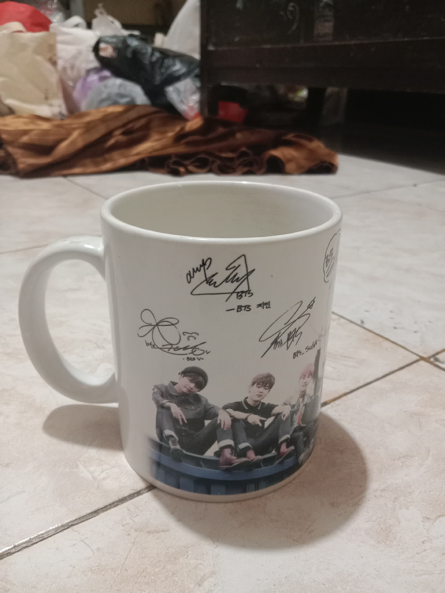 The image shows a white ceramic mug with designs on it, including what appear to be autographs and pictures of K-pop group BTS members. The mug is placed on a tiled floor. In the background, there are some cloth items and what looks like discarded plastic bags, suggesting a somewhat cluttered environment. The lighting suggests an indoor setting, possibly a home. There is no information in the image to suggest the location is the city of Madiun, Indonesia.
