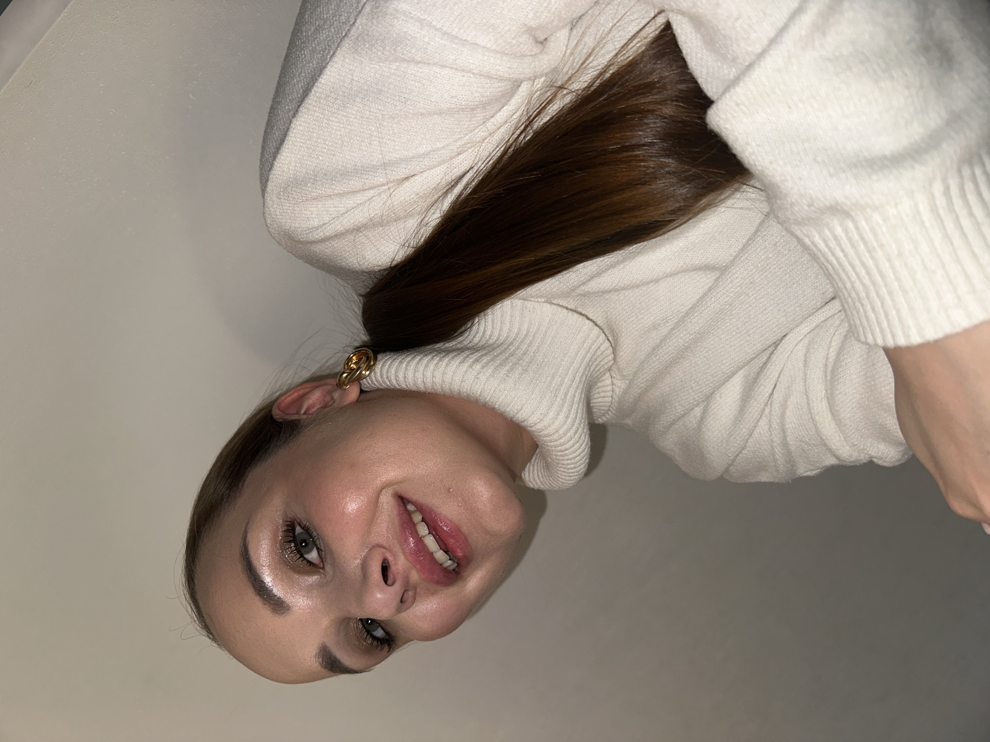 A young woman with fair skin and light brown hair is wearing a white turtleneck sweater and a gold earring. She is looking at the camera with a slight smile. The background is a plain white wall. The location is not specified in the image.