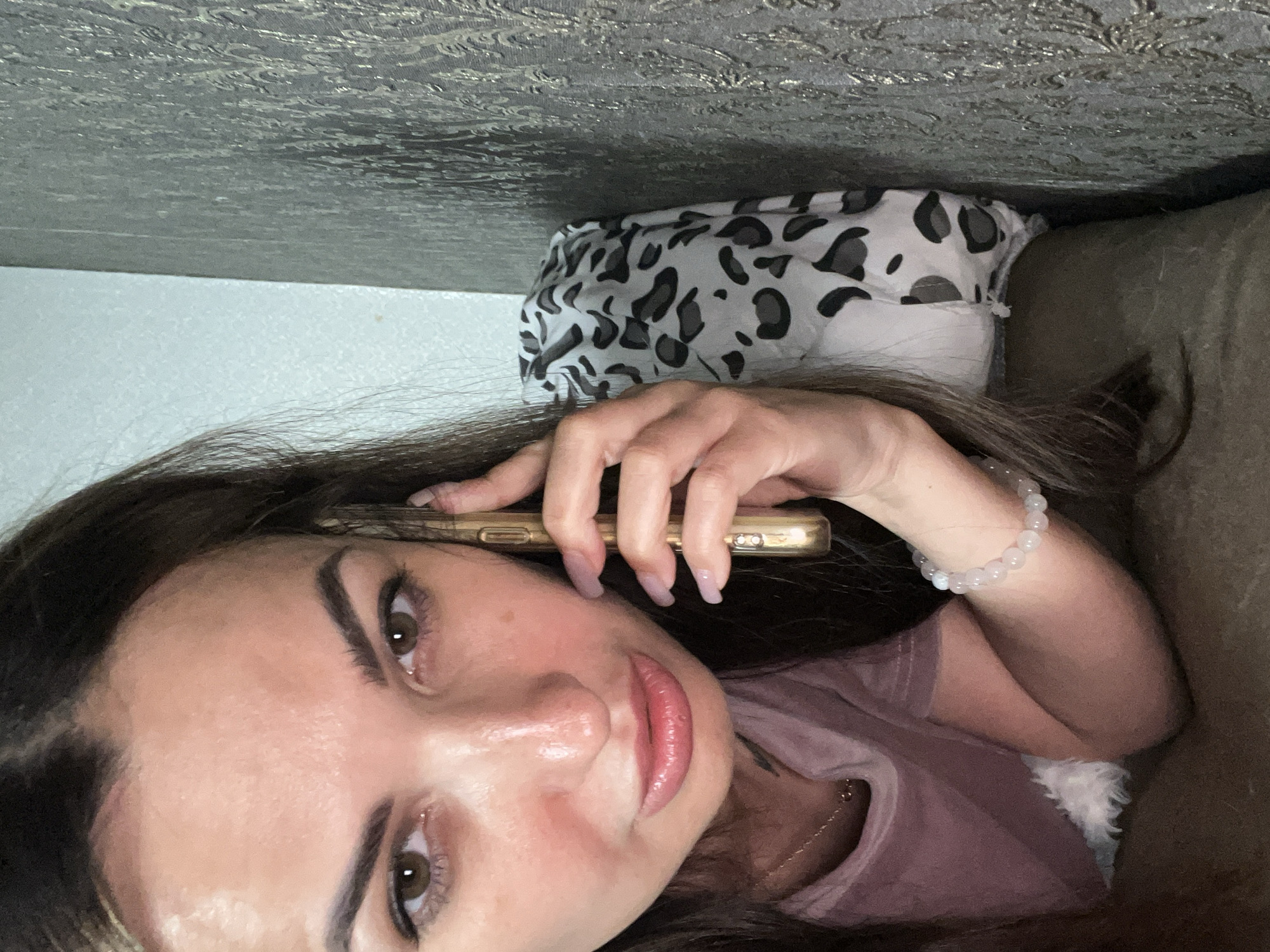 The media file is a close-up, selfie-style photograph of a woman holding a gold-colored smartphone to her ear. She has long, dark hair and is looking towards the camera with a neutral expression. Her eyes are brown, and she is wearing makeup, including mascara and lipstick. She is also wearing a light pink t-shirt and a beaded bracelet on her left wrist. The background of the image is dark and appears to be a couch or bed. There is a pillow with a black and white leopard print behind her. The lighting suggests it might be indoors, possibly at night or during the day with artificial light. There is no visible text in the image. The overall mood of the image is casual and intimate.
