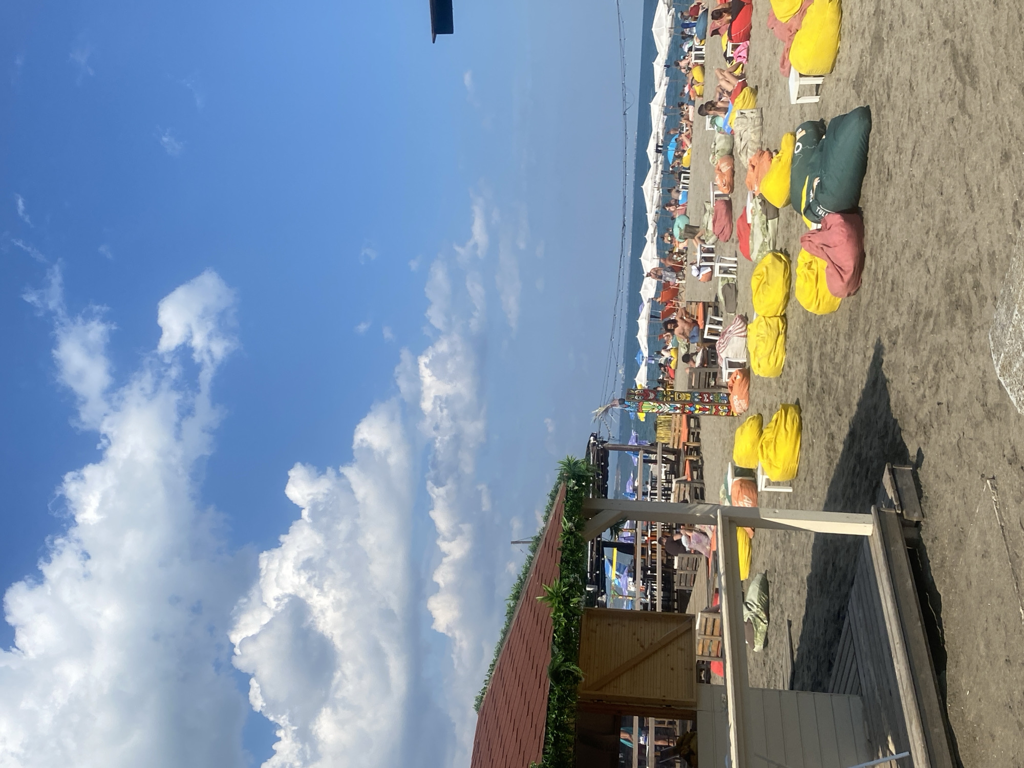 The image depicts a busy beach scene on a sunny day in Burgas, Bulgaria. The foreground features a wooden structure with a red roof, possibly a kiosk or bar, decorated with artificial greenery. Several large, brightly colored beanbag chairs are scattered on the dark sand, some occupied by people. In the midground, rows of white umbrellas are set up on the beach, with numerous individuals relaxing or socializing underneath them. A body of water, likely the Black Sea, stretches to the horizon, meeting a bright blue sky dotted with fluffy white clouds. The overall atmosphere is one of leisure and recreation.