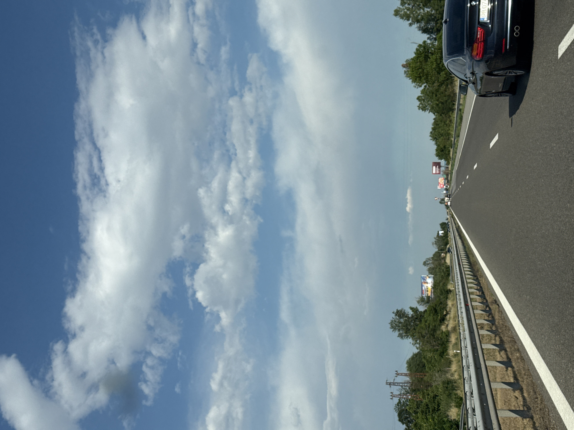 The image shows a view from a car traveling on a highway near Kalugerovo, Bulgaria. The highway has multiple lanes and a metal guardrail separates it from the grassy shoulder. On the left side of the highway, there are trees, utility poles, and billboards. On the right side, a black BMW sedan is visible, traveling in the same direction as the camera car. The sky is blue with scattered white clouds. The scene is captured during daylight.