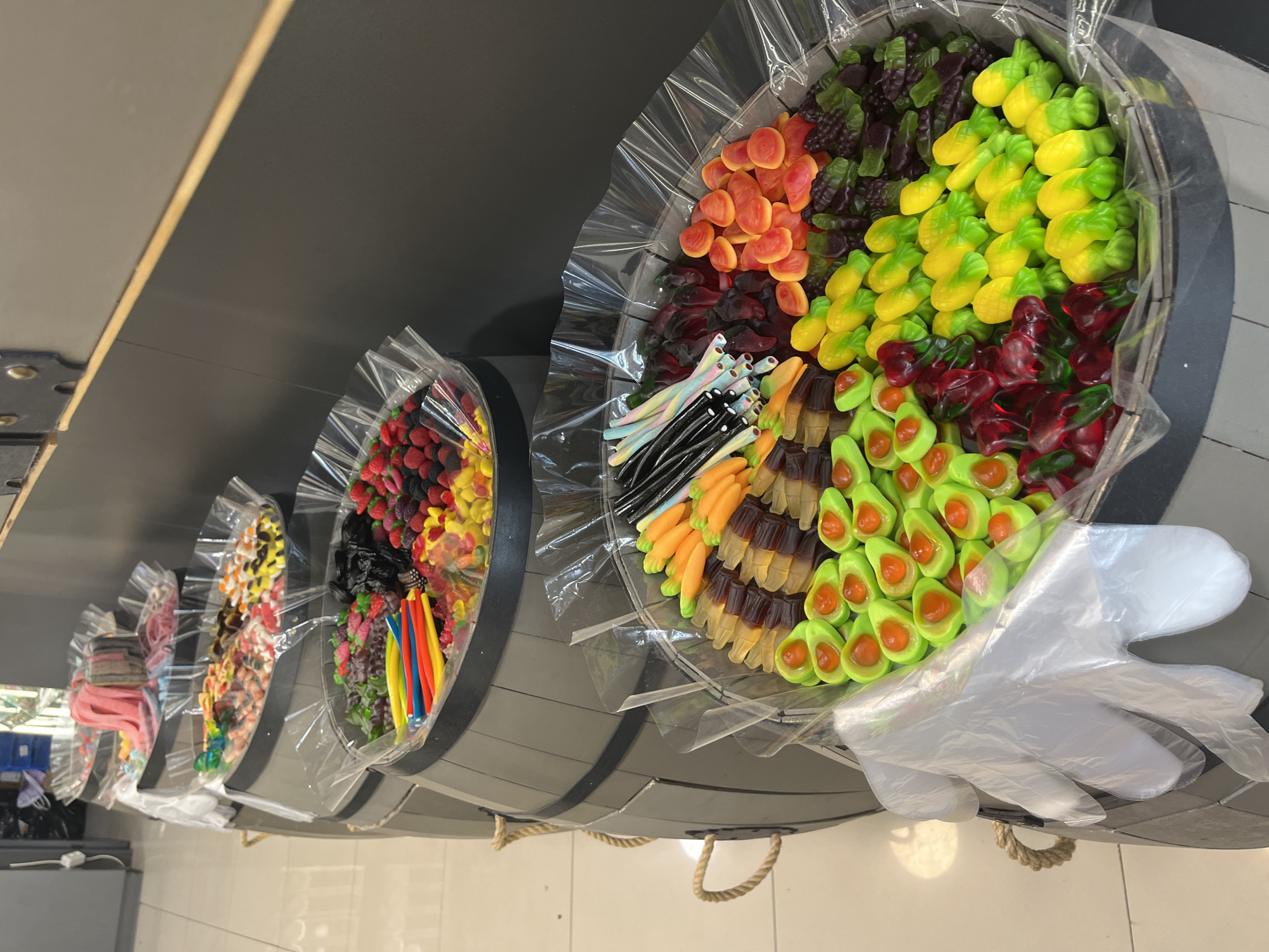 This media file depicts a display of gummy candies in a store. The candies are arranged in what appear to be wooden barrel-shaped containers, each sectioned off and covered with clear plastic packaging. The candies themselves are diverse in shape and color, including gummy pineapples, cherries, avocados, carrots, and other assorted fruit shapes. Some sections contain licorice-like candies and colorful striped candy sticks. In the foreground, a pair of clear plastic gloves is visible, suggesting that customers can use them to handle the candies. The overall impression is one of a vibrant and abundant candy selection in a retail environment. There is no visible text in the image. The lighting suggests an indoor setting, likely a candy store or a section of a larger grocery store.