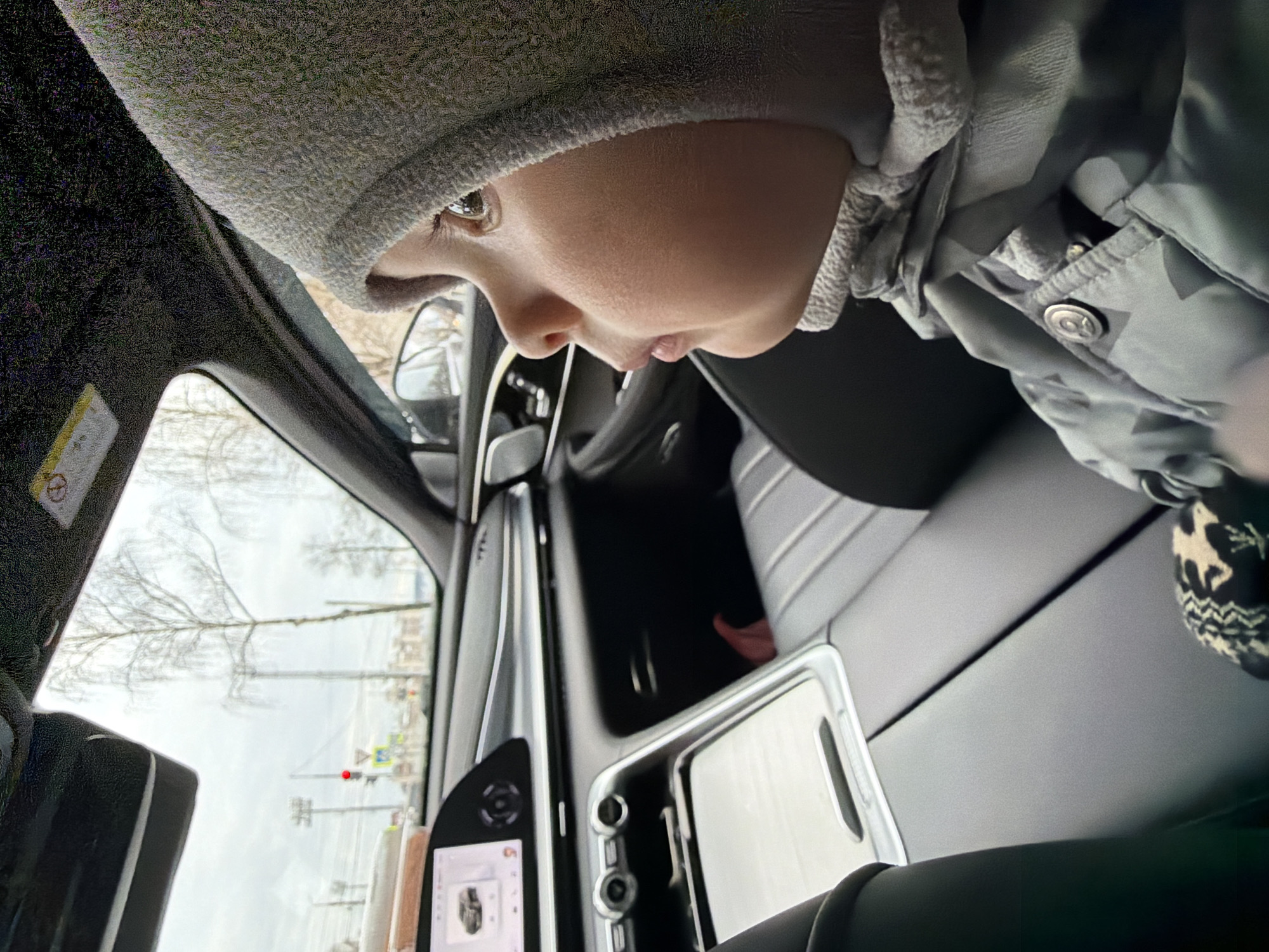 A close-up, Dutch-angle shot captures a young child from the chest up, situated within the interior of a modern car. The child is wearing a grey knitted hat and a grey/blue winter jacket, suggesting cool or cold weather. Their profile is visible, with light brown or hazel eyes looking downwards and to the left, likely observing something inside the car or out the window.
The car's interior features a black dashboard, a central console with a screen displaying faint information, and part of a steering wheel. A smartphone or tablet in a clear case is mounted on the console. Through the side window, a grey, overcast sky is visible, along with bare trees and a street scene. Outdoor elements include a road or pavement, power lines, and a street sign with a red circular light, possibly a traffic light or pedestrian crossing indicator. A blurred car side mirror is also visible.
In the bottom right corner, a child's foot or ankle, clad in a black and white patterned sock or boot adorned with reindeer-like figures, is partially visible. A yellow warning label is present on the car's interior pillar near the windshield, but its text is unreadable. The scene depicts a quiet, daytime moment in Votkinsk, Russia, during a cold season like late autumn or winter, with the child engaged in observation from inside the vehicle.
