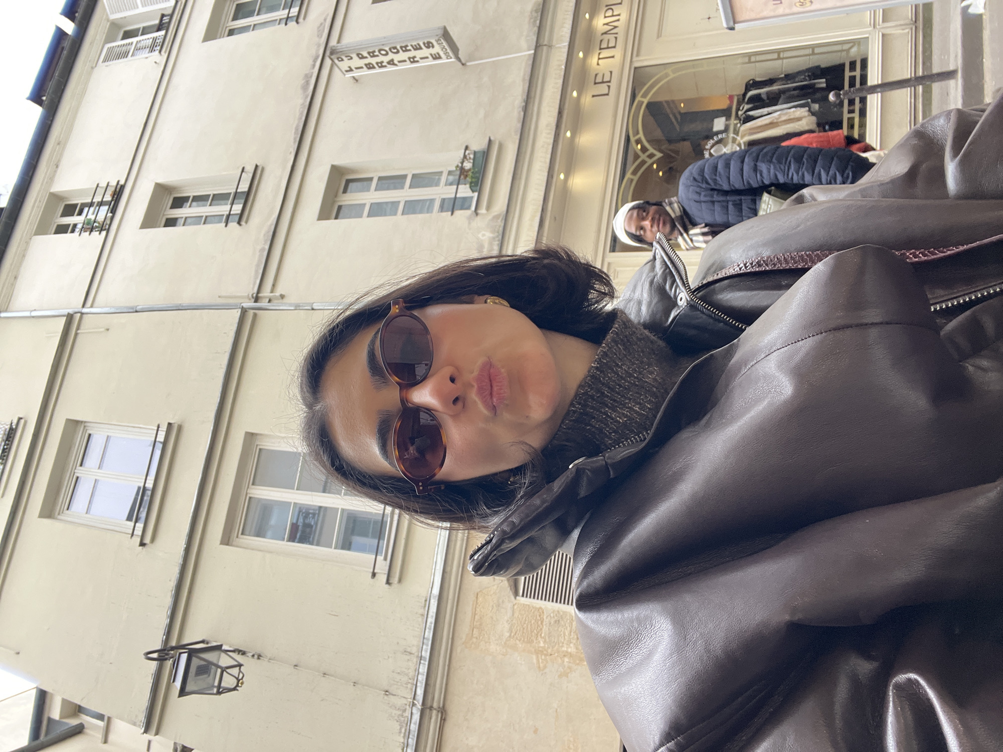A woman wearing sunglasses and a brown leather jacket is taking a selfie. Behind her is a light yellow building with multiple windows and a street lamp. The building's sign reads "Le Temple," indicating the location is in Paris, France. In the background, another person is wearing a white beanie and a dark jacket, sitting at what appears to be an outdoor seating area.