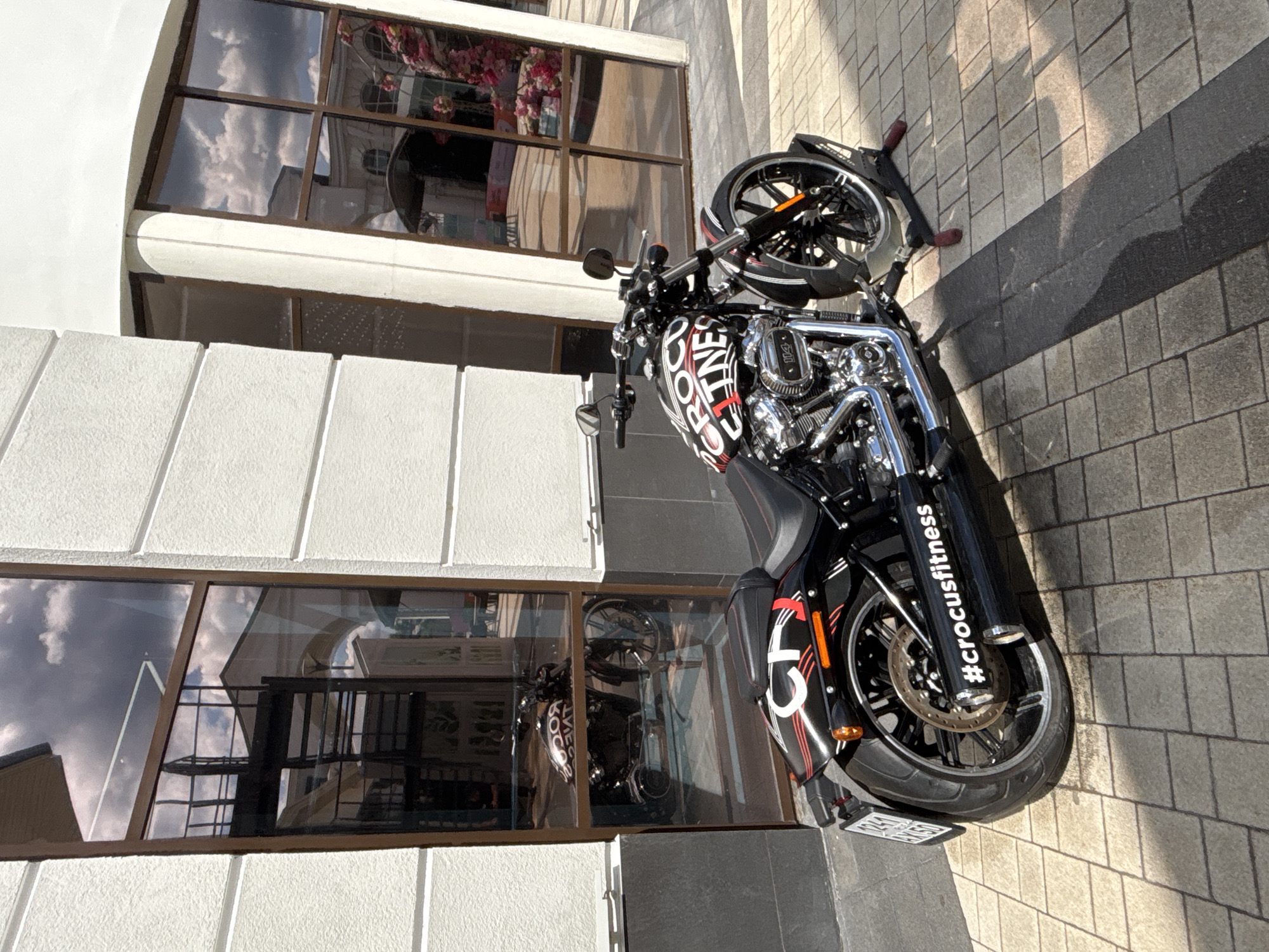 A black Harley-Davidson motorcycle is parked on a paved walkway outside a building with large reflective windows. The motorcycle has "CROCCFITNESS" in white lettering on its fuel tank and "#crocusfitness" on its exhaust pipe. The windows reflect the sky, clouds, and parts of the building's interior, including what appears to be a staircase and possibly some plants. The building is modern in design with white walls and brown window frames. The walkway is made of interlocking square pavers, and shadows of the motorcycle and the building fall across it. The scene appears to be set in daylight.