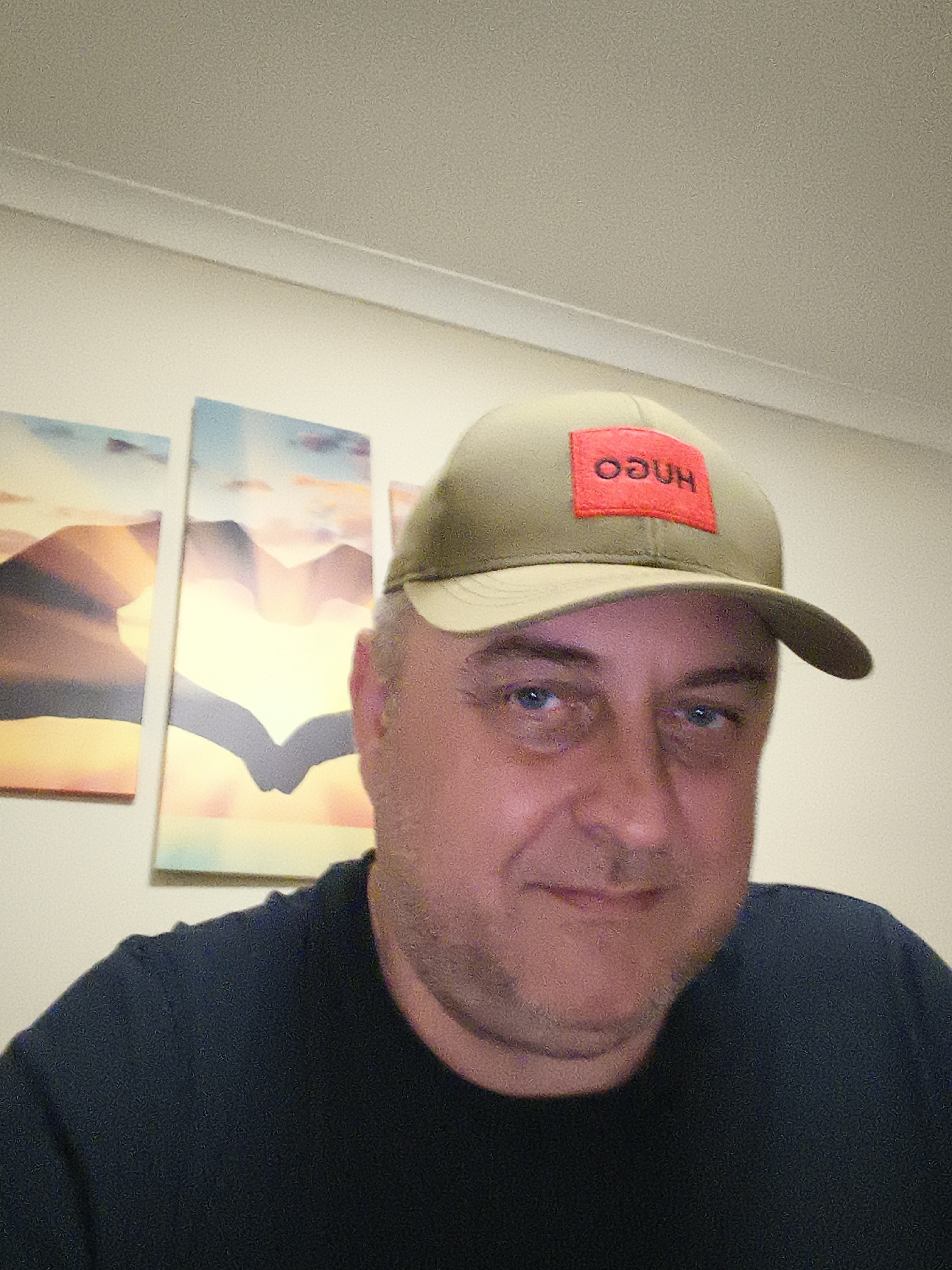 The image captures a head-and-shoulders portrait of a middle-aged man in an indoor setting, likely a residence in Huntingdonshire, United Kingdom. He has light skin, blue eyes, and visible facial stubble, and is looking directly at the camera with a calm, slightly smiling expression.
The man is wearing a dark blue t-shirt or crewneck shirt. On his head, he sports an olive green baseball cap. The front of the cap features a prominent red rectangular patch with white text. The text, "hugo," is upside down from the viewer's perspective, appearing as "oƃnh."
In the background, a light-colored wall is visible, on which three rectangular art prints or canvases are mounted. These artworks depict abstract or stylized landscape scenes, possibly sunrises or sunsets over mountains, characterized by soft blends of orange, yellow, and blue hues. A white ceiling with a molding strip is visible at the top of the frame. The lighting in the image appears to be artificial and diffused, indicating an indoor environment, possibly in the evening or night. The man's relaxed pose suggests a casual moment, perhaps a selfie or a candid photograph.