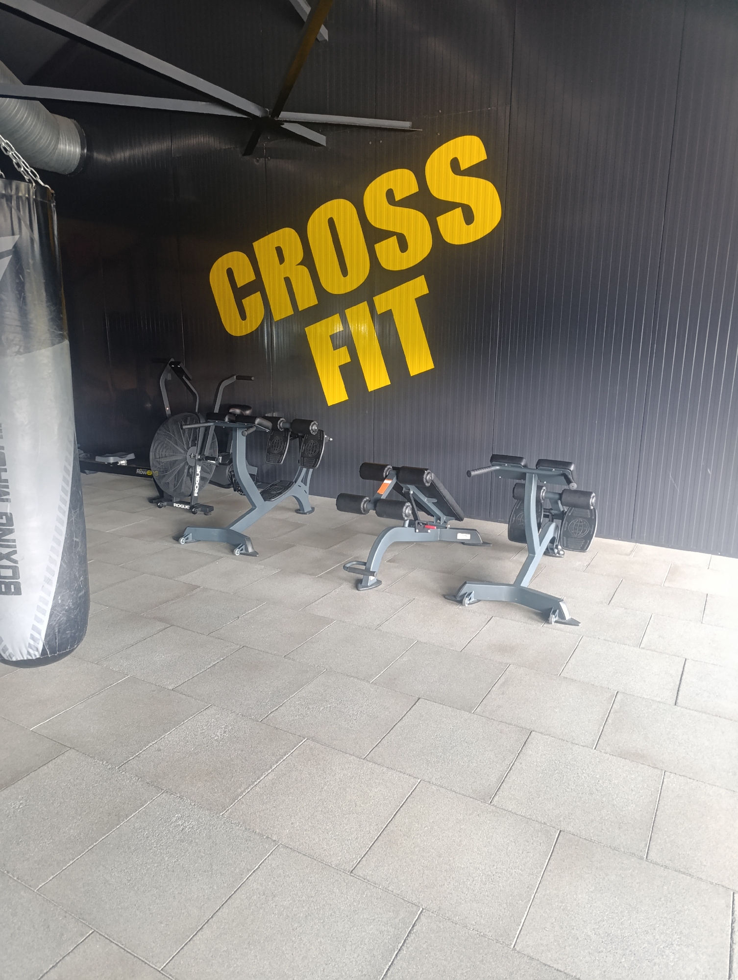 The image depicts the interior of a gym, likely a CrossFit facility, located in Zlatopil, Ukraine. A large, grey punching bag branded "BOXING MASTER" is visible on the left side of the frame. In the background, a wall painted black features large yellow lettering that reads "CROSS FIT". Several pieces of gym equipment are present, including a Rogue Air Bike and two abdominal crunch benches. The floor is covered with light grey, rectangular tiles. The environment appears clean and well-maintained, with industrial-style lighting and ventilation visible on the ceiling. No people are present in the image.