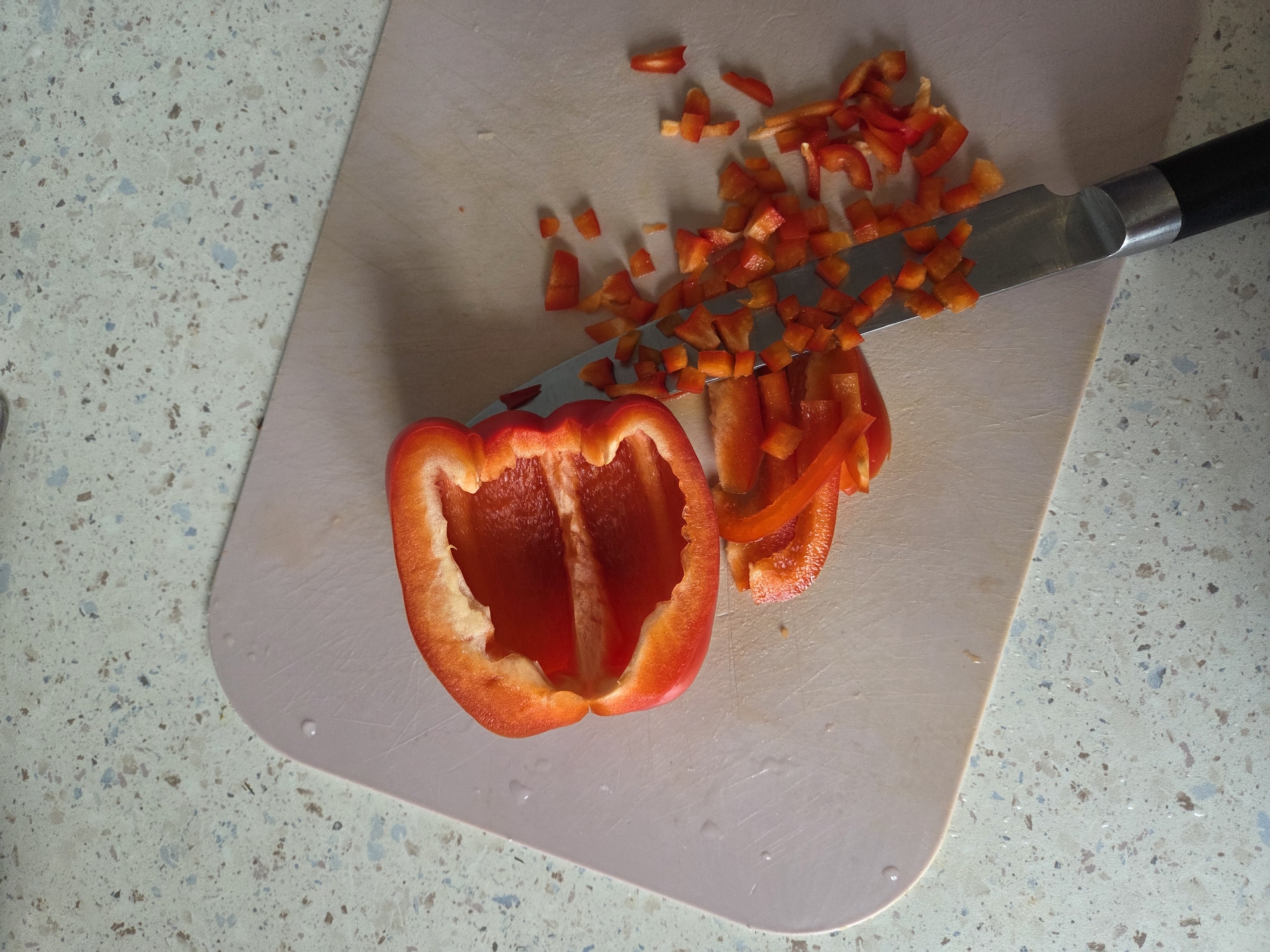 The image shows a red bell pepper, partially chopped, on a light pink cutting board. A silver knife with a black handle is positioned over the cutting board with its blade resting on the chopped pepper pieces. The cutting board is placed on a speckled, light-colored countertop. The location, Birobidzhan, Russia, is not evident from the visual information provided.