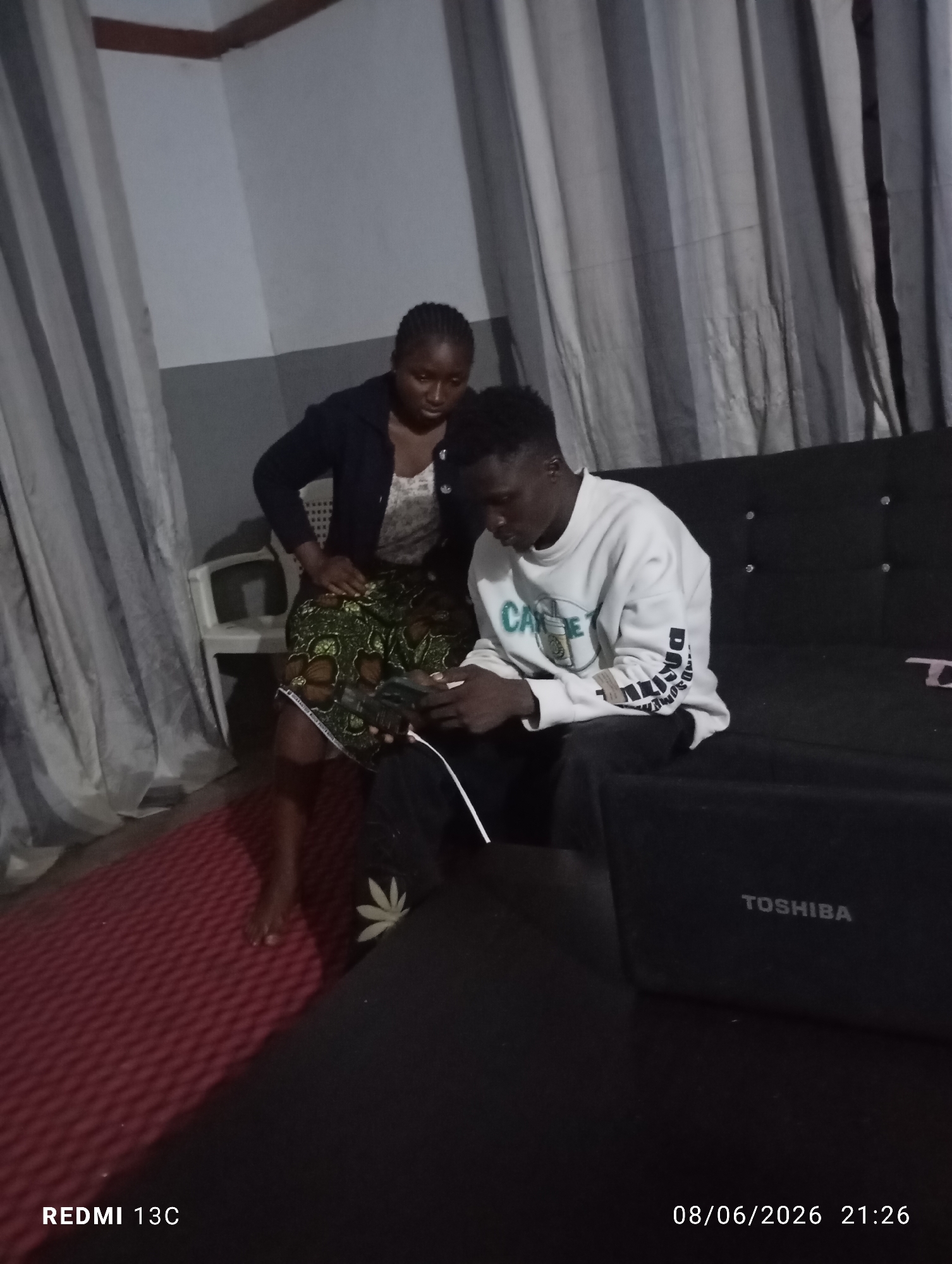 A medium shot captures two individuals, a woman and a man, in what appears to be a living room in Jos, Nigeria. The woman, positioned on the left, is standing and looking down, wearing a dark jacket over a patterned top and a patterned skirt. The man is seated on the right, engrossed in a mobile phone he is holding. He is dressed in a white sweatshirt with lettering on the front and sleeves, and dark trousers. A black sofa is the primary seating furniture, with a laptop open on its surface, displaying the "TOSHIBA" logo. To the left of the sofa, a red textured mat covers the floor. The background features a grey wall and curtains. The scene is dimly lit, suggesting it might be evening, as indicated by the timestamp "08/06/2026 21:26" visible in the lower right corner, along with "REDMI 13C" in the lower left.