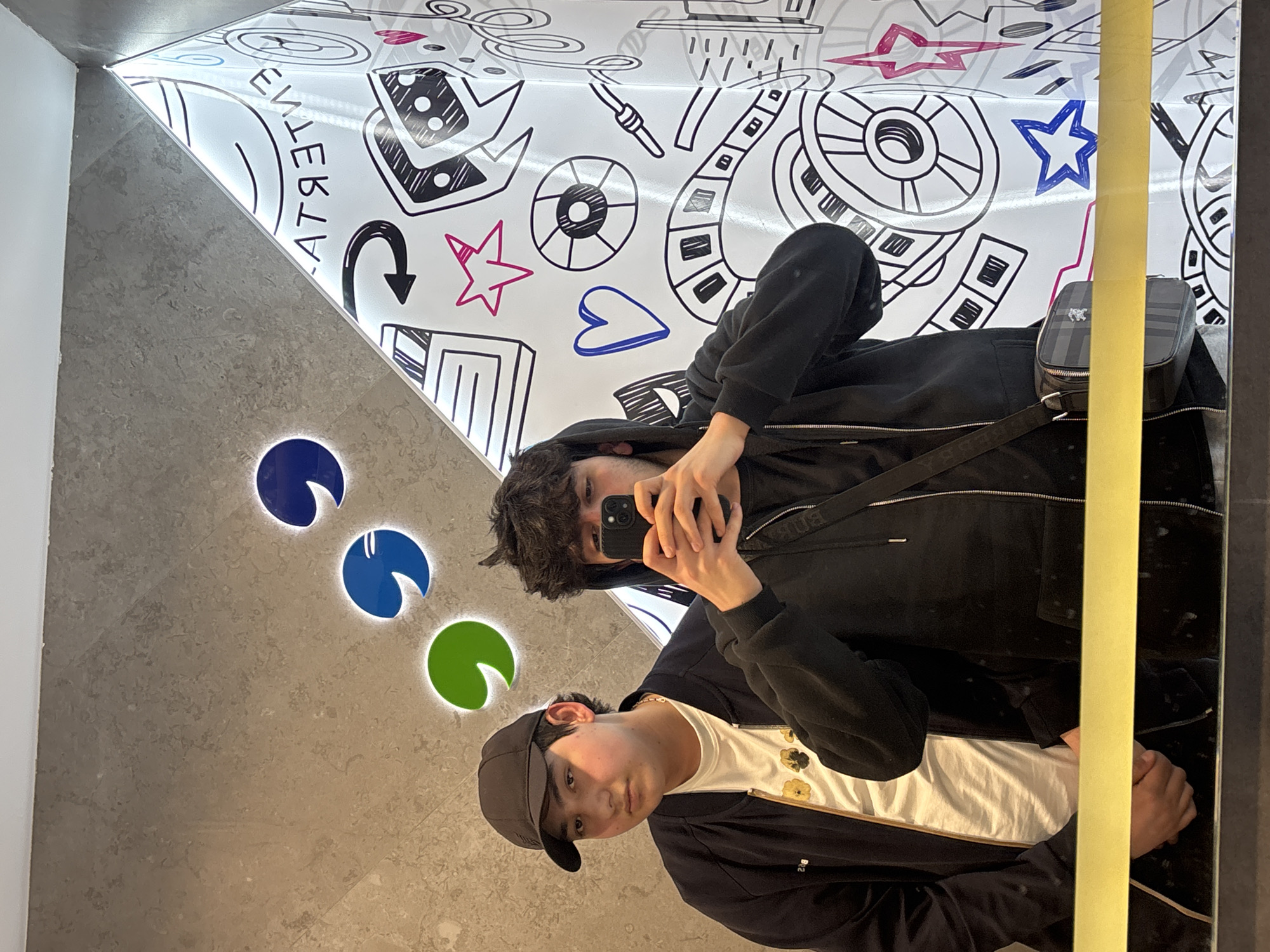 A photograph shows two young men taking a selfie in a mirror within a building in Yerevan, Armenia. The man closest to the camera wears a black baseball cap, a black jacket, and a white T-shirt with a floral pattern. The other man, positioned higher in the frame, wears a black hooded sweatshirt and holds a phone to take the picture. Both men are reflected in the mirror.
The background behind the mirror to the left is a wall with three illuminated circular designs in blue and green. Above the men in the mirror is a white surface adorned with various black and white illustrations, including musical notes, stars, hearts, and abstract shapes. A vertical yellow strip is visible along the right edge of the mirror.