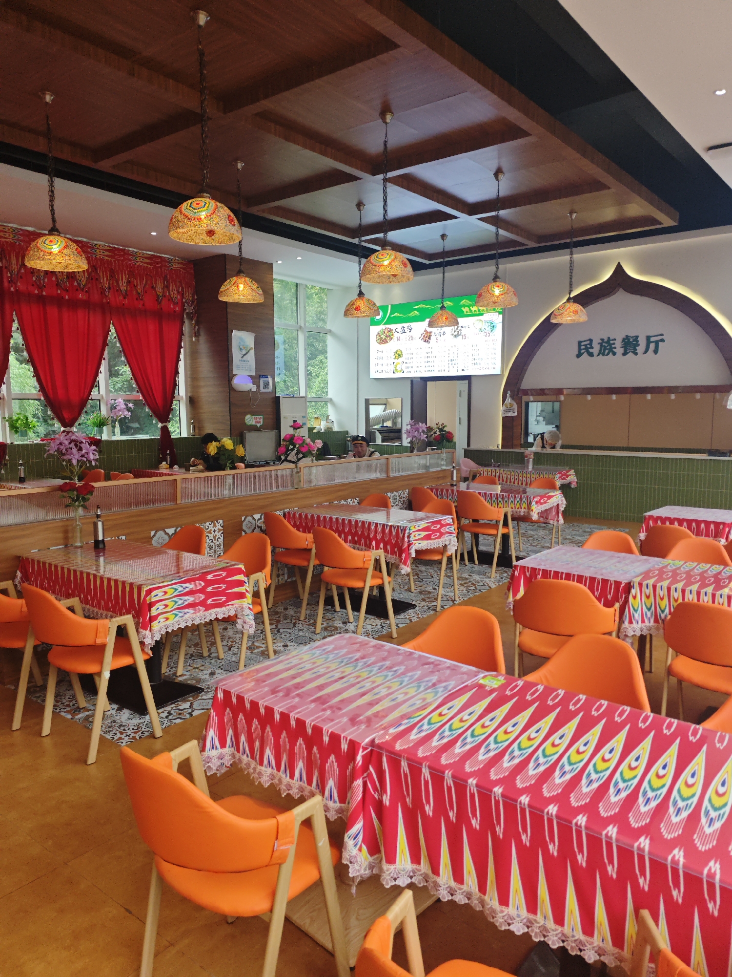This is an interior shot of a restaurant in the Chongqing High-tech Zone, China, featuring vibrant orange chairs arranged around tables covered in red tablecloths with a colorful, patterned design. The ceiling is adorned with wooden panels and decorative pendant lights with mosaic shades. Red curtains hang on the left side of the image, and large windows in the background reveal greenery outside. A menu board with Chinese characters is visible on the wall above the counter area, and there is a hint of activity behind the counter with a few individuals present. The overall ambiance suggests a lively and ethnic dining establishment.