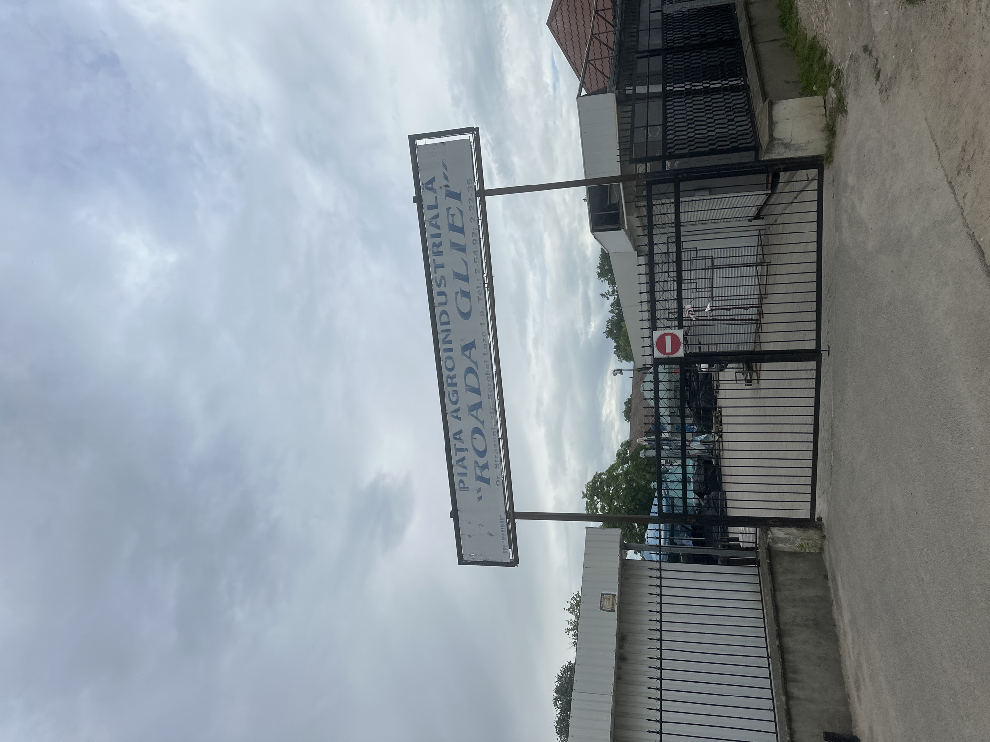 This is a photograph of the entrance to "PIATA AGROINDUSTRIALĂ ROADA GLIEI" in Strășeni, Moldova. A large white sign with blue text, mounted on a pole, displays the name of the market and its address: Str. Serghei Lazo 1-a. Below the name, contact telephone numbers are listed. The sign is tilted, with the text running vertically.
The entrance itself is secured by a black metal gate, which is partially open, allowing a view into the market. Inside the gate, several vehicles, possibly cars and vans, are visible parked. The setting appears to be an outdoor market or a commercial area.
To the right of the gate, a concrete pathway leads away from the entrance. A red and white circular sign with a horizontal bar, indicating "no entry" or a prohibition, is affixed to the gate.
The sky above is overcast with significant cloud cover, suggesting a cloudy or potentially rainy day. The overall lighting is diffused, indicating it might be daytime, but the exact time is not discernible. The photograph captures a moment of everyday activity at a commercial hub in Strășeni.