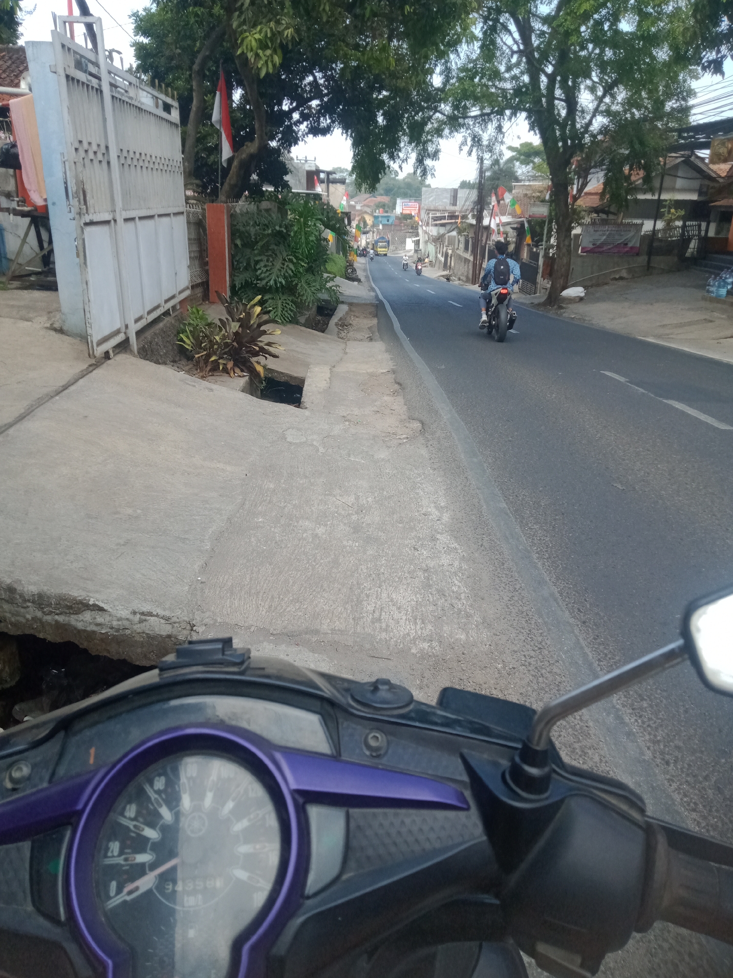 The image shows a street scene in Sumedang, Indonesia, as viewed from the perspective of a motorcycle rider. In the foreground, the motorcycle's dashboard is visible, featuring a speedometer with a purple trim and the "Yamaha" logo. The road ahead is asphalt with a white line marking the lane.
To the left of the road, a concrete sidewalk runs alongside a white metal gate and dense green foliage. The gate appears to be made of vertical bars. Behind the gate, a tall tree with green leaves is prominent. Further down the street, several buildings line the right side, some with decorative flags.
On the road, a person is riding a motorcycle, facing away from the viewer. The rider is wearing a blue shirt with a backpack. Other motorcycles and a yellow truck are visible further in the distance, indicating traffic. The overall environment suggests a moderately urban or suburban area with a mix of residential and commercial structures, typical of a developing city. The time of day appears to be daytime, with natural light illuminating the scene.