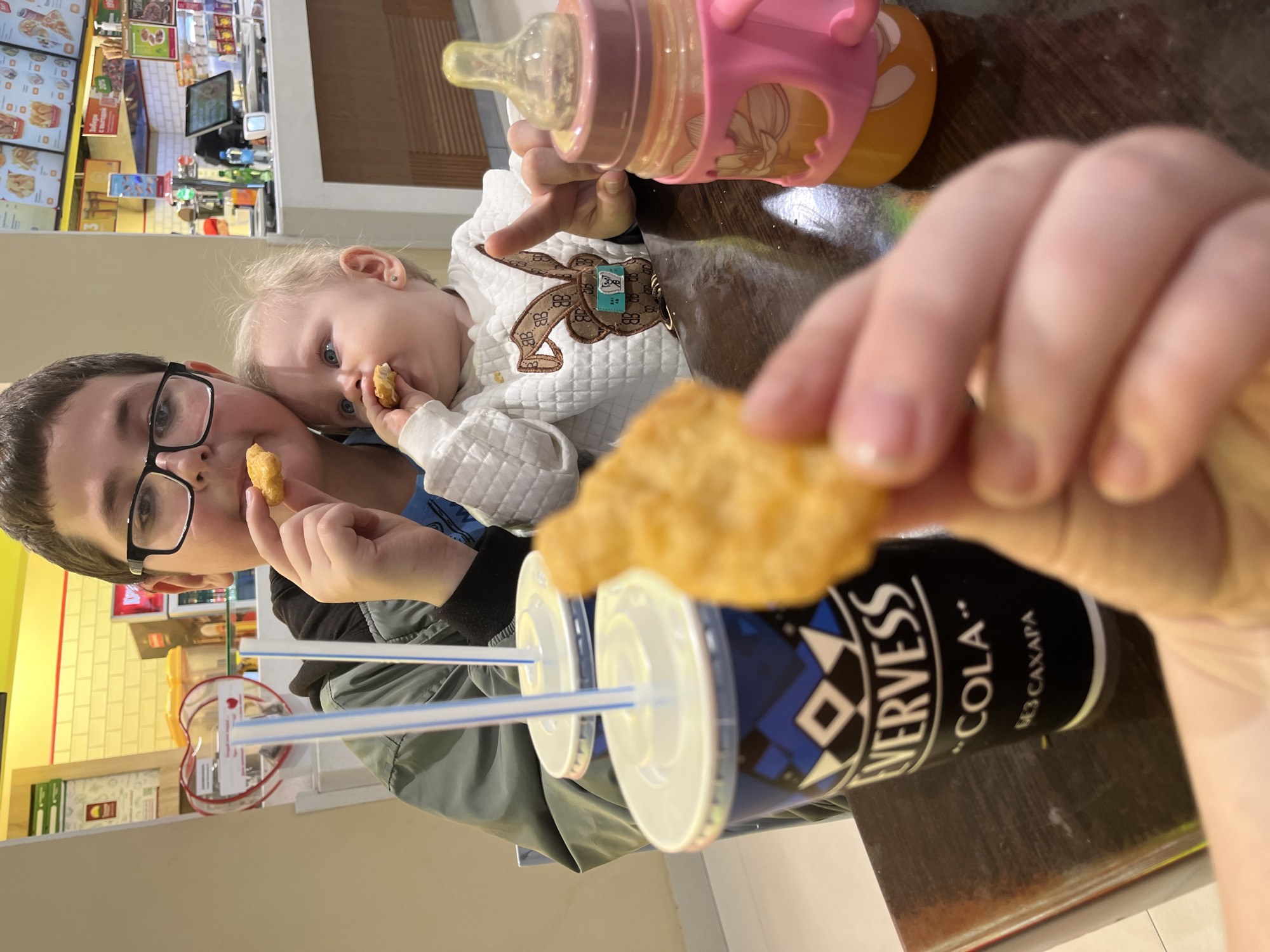 Here is a detailed description of the image:
**People:** The image features a young man wearing glasses and a baby. Both are holding and apparently eating chicken nuggets.
**Subjects:** The subjects are the two people having a meal together.
**Objects:** The image contains two cups of "Evervess Cola," a baby bottle with juice, and chicken nuggets. The baby's clothing has a decorative patch.
**Actions:** The people are eating, and the image captures them in a moment of enjoying a meal together.
**Interactions:** The interaction is between the two people, where the older individual appears to be helping the younger one eat.
**Environment:** The scene is set in what appears to be a fast-food restaurant. Based on the "Evervess Cola" branding, the location is in Russia, presumably in the city of Sochi. This conclusion is based on the use of Russian script ("БЕЗ САХАРА" meaning "without sugar") on the cola cups. The restaurant setting is evident from the visible menus, the interior design, and the general ambiance in the background.