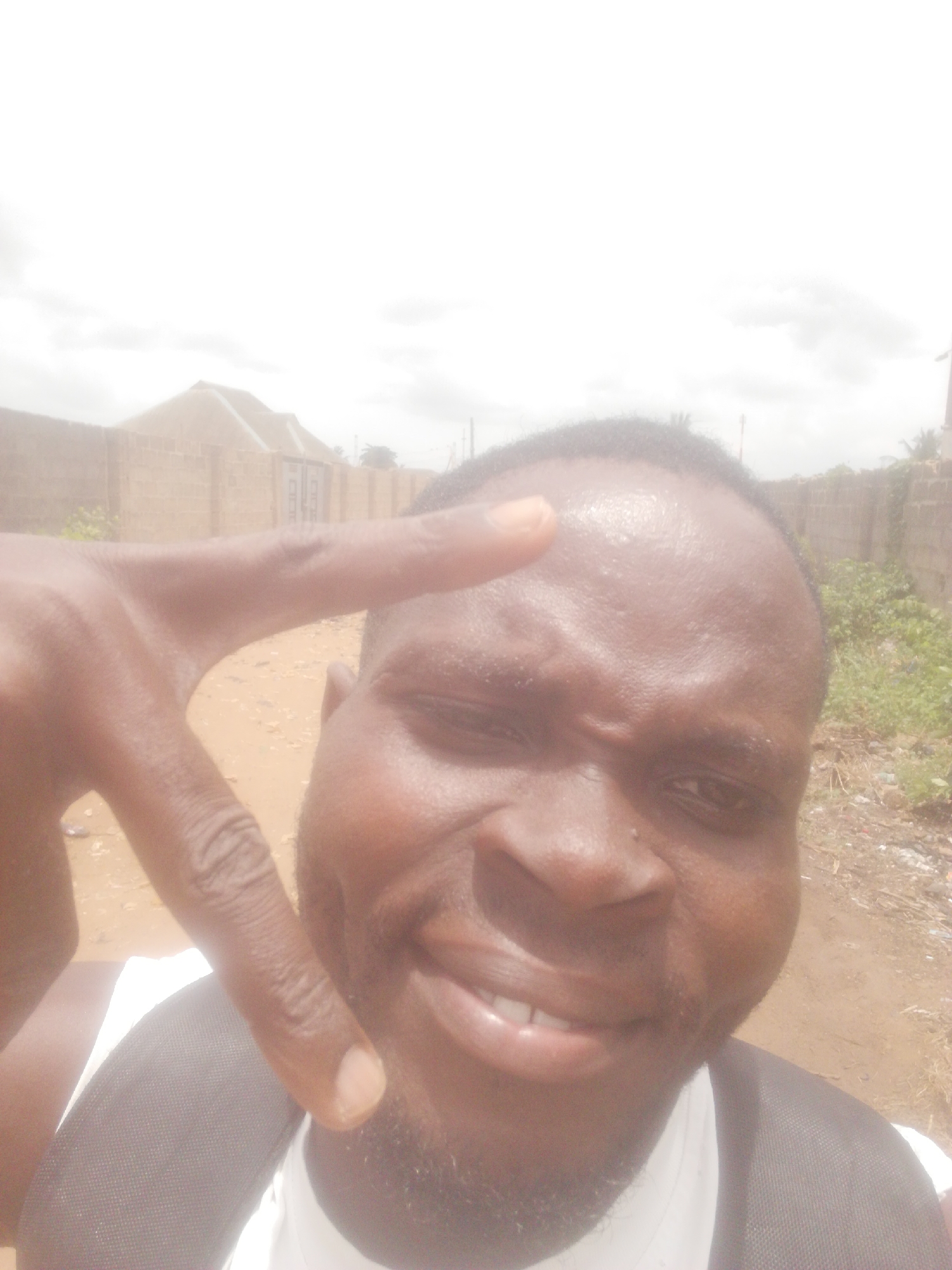 A close-up selfie depicts a Black man with short hair and a beard, holding up a peace sign with his right hand. The man is smiling and his face appears moist. He is wearing a dark colored backpack over a white shirt.
The background shows a dusty, sandy area, possibly a dirt road or open ground. A partially constructed building with a brown roof is visible behind a brown brick wall in the distance. Some green vegetation is sparsely scattered along the wall. The sky is overcast. The location appears to be in a semi-urban environment.
