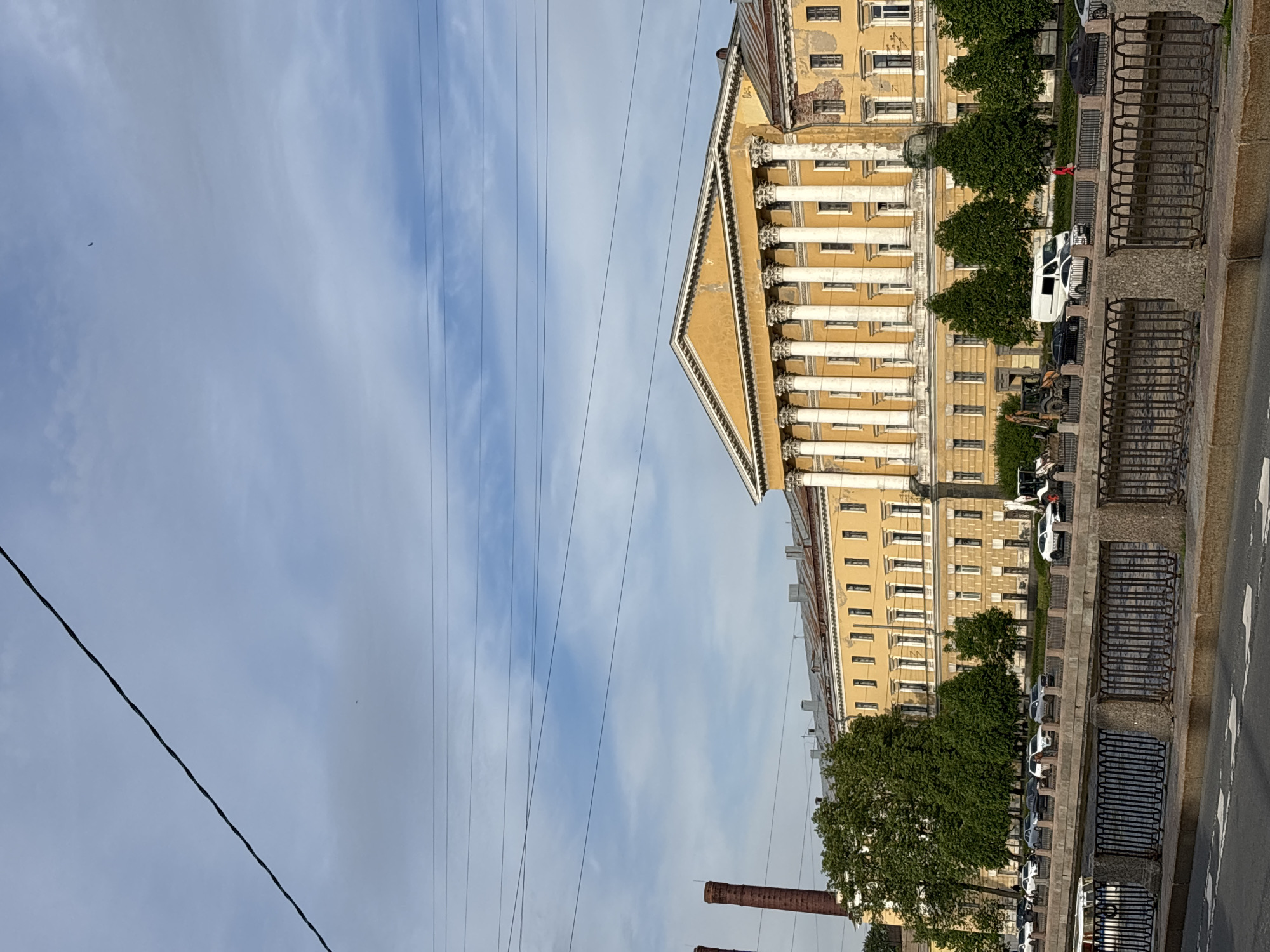 An urban landscape in Saint Petersburg, Russia, features a grand, multi-story building with distinct classical architectural elements. The building, painted in a warm yellow hue, boasts numerous white columns and a prominent triangular pediment at its central peak, adorned with decorative details. Rows of windows are visible across its facade, which shows signs of age.
In front of the building, a tree-lined street or embankment runs parallel, with several vehicles, including white vans and cars, parked along the curb. A sturdy, dark metal railing, likely part of an embankment or bridge structure, separates the sidewalk from an adjacent roadway or canal bank. A person in red is visible on the sidewalk in one of the provided crops.
The sky above is partly cloudy, with streaks of white clouds against a pale blue background. Several thin power lines or wires traverse the upper part of the image. A small, dark, elongated object, possibly an aircraft, is faintly visible in the upper sky. In the lower left corner, two brick chimneys rise, indicating other structures in the vicinity. The scene is illuminated by daylight, suggesting daytime conditions. No discernible text is visible.