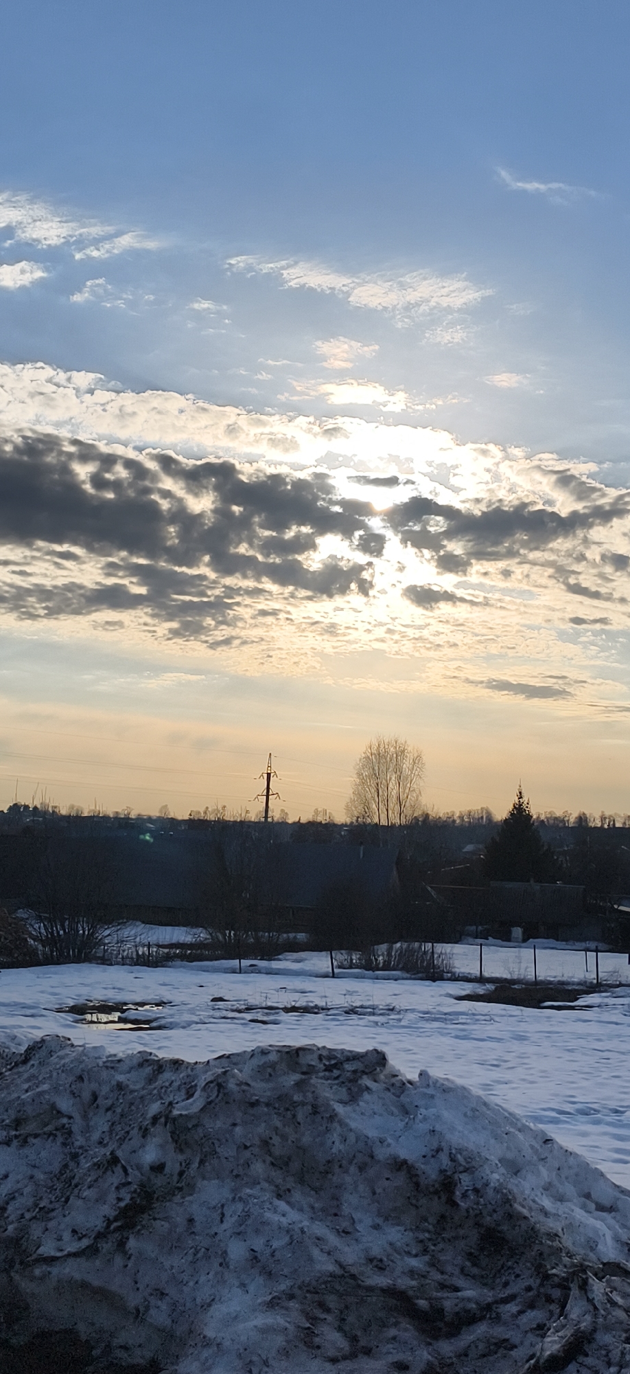 The image shows a winter landscape in Фоки, Russia. Patches of snow cover the ground, with a large pile of dirty snow in the foreground. Puddles of water are visible amidst the remaining snow. In the midground, there are houses and buildings typical of a rural setting. Power lines are present, running across the horizon. The sky is mostly blue with scattered clouds, and the sun is partially obscured by the clouds, creating a diffused light. The overall impression is of a cold, overcast day in a village.