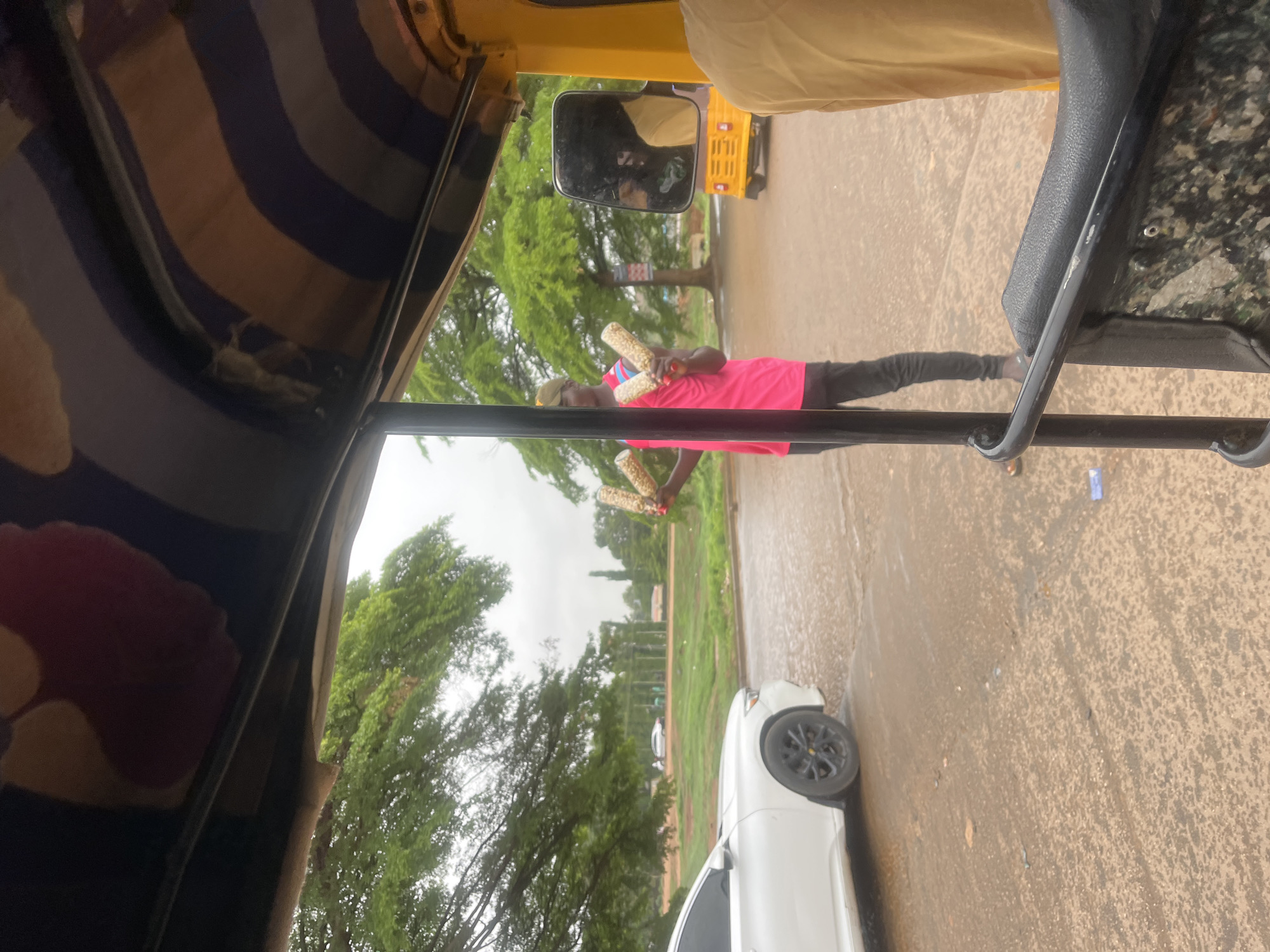 A man in a pink shirt stands on the side of a road holding what appears to be corn on the cob in both hands, seemingly offering it for sale. A white car is parked nearby. The scene is viewed from the inside of a three-wheeled vehicle, possibly an auto rickshaw. The location is identified as Galadimawa Village, Nigeria. The road is paved and wet, possibly from recent rain. Trees and greenery are visible in the background.
