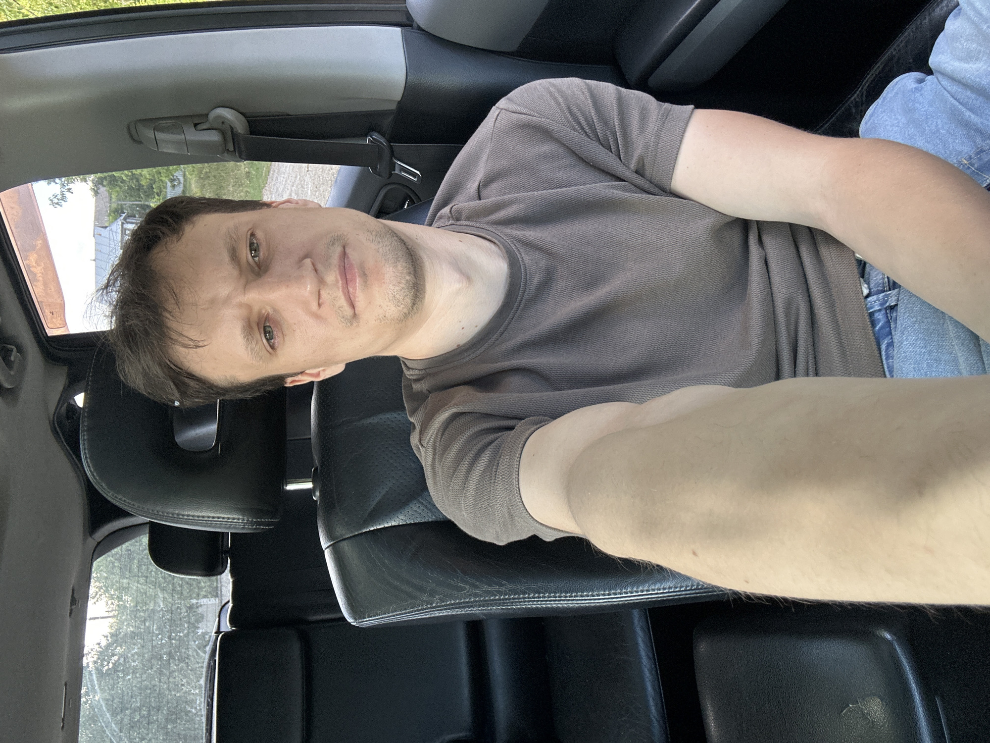 A man is taking a selfie in the driver's seat of a car. He is wearing a brown t-shirt and blue jeans. The car appears to be parked on a dirt road, with houses and trees visible in the background. The location is Makiv, Ukraine. The man's arm is extended, holding the camera. The interior of the car is dark, with leather seats visible. Sunlight is coming through the windows.
