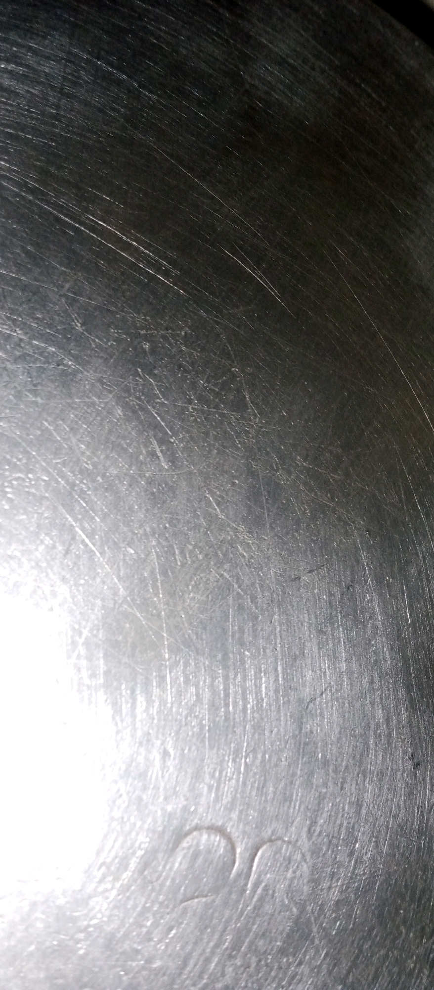 The image displays a close-up, high-angle shot of a scratched, metallic surface. The surface has a brushed or polished finish, with numerous fine, parallel scratches and some deeper gouges. Light reflects off the surface, creating bright highlights and obscuring some details. In the lower portion of the image, there are two faint, curved indentations that resemble stylized Arabic numerals or symbols. The overall impression is of a worn or aged metal object. There is no discernible urban environment or location information visible in the image itself.