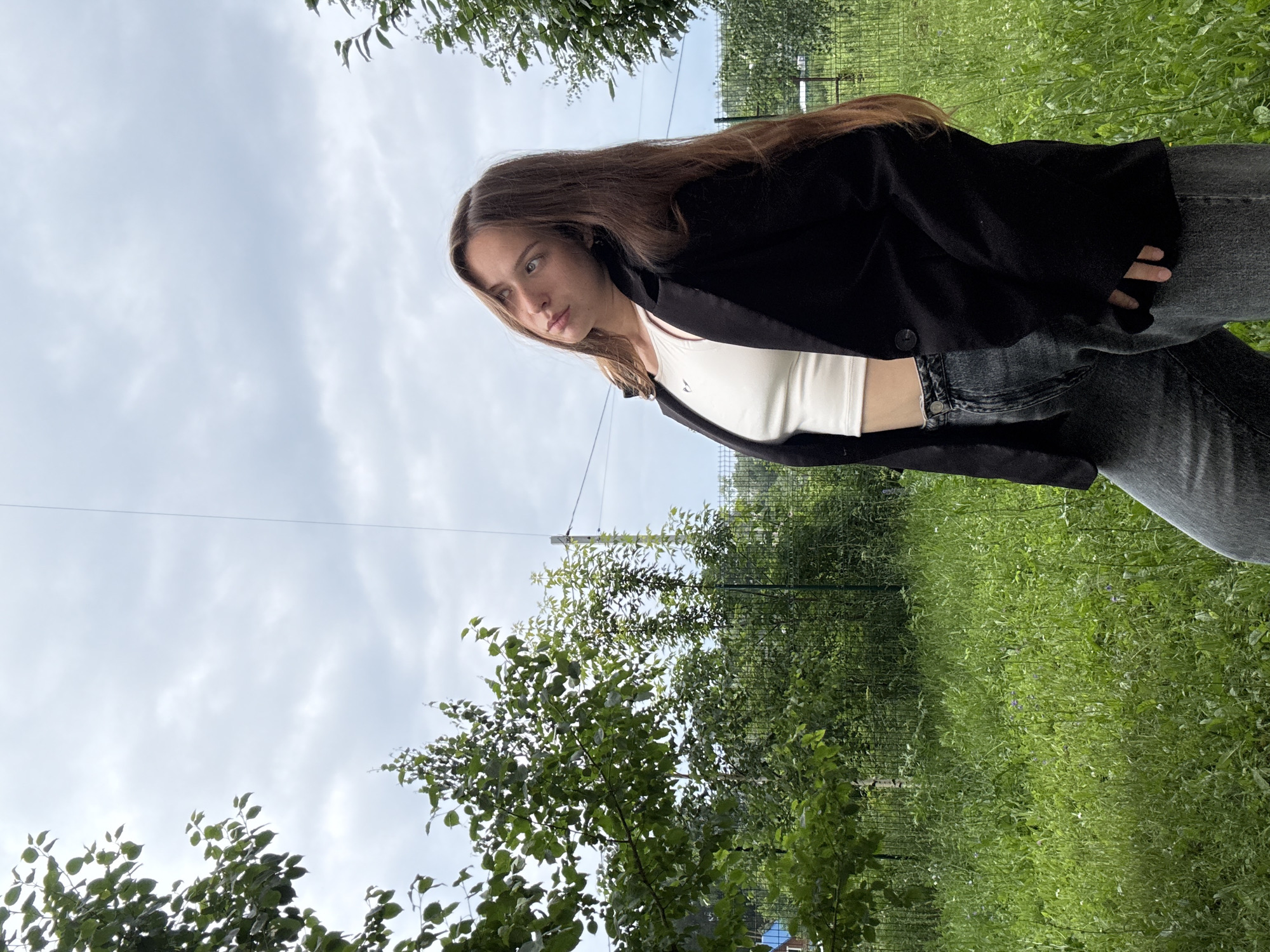 A young woman with long, brown hair is shown from the chest up, looking down and to her left. She is wearing a black blazer over a white tank top and dark jeans. Her expression appears thoughtful or contemplative. The background consists of a cloudy sky above and a dense green grassy area with trees on the right. A wire or cable is visible diagonally across the upper portion of the image. The scene is outdoors, with natural light suggesting it is daytime. The overall mood is casual and relaxed.