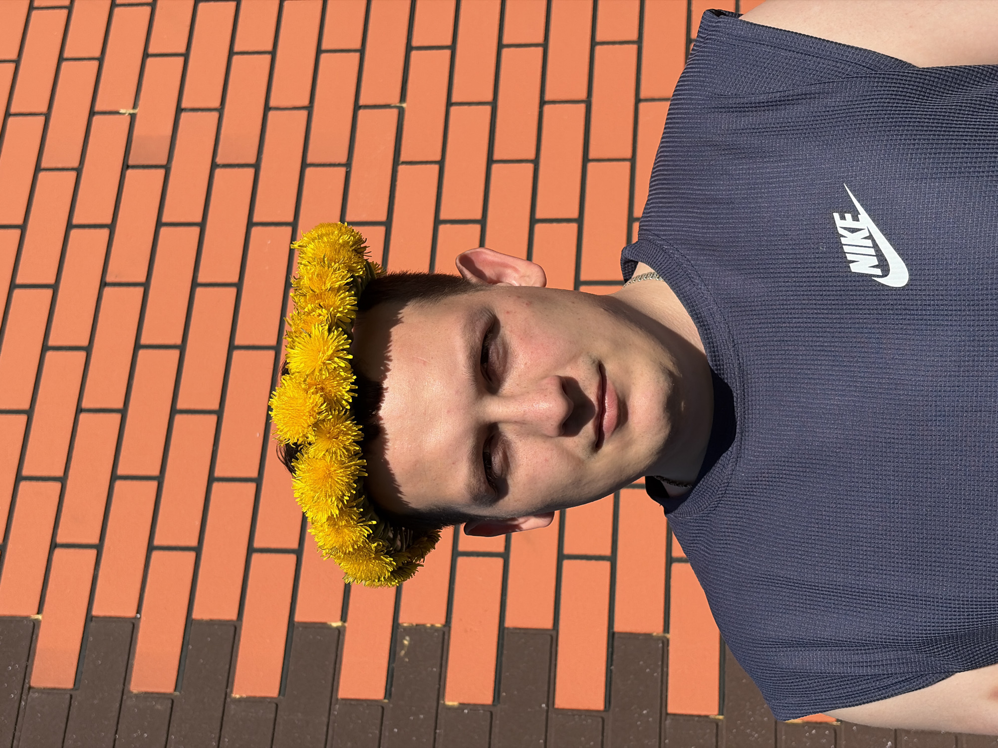 A person wearing a yellow dandelion crown is positioned on a brick surface. The person has short, dark hair and is wearing a navy blue Nike t-shirt. The background consists of light orange and dark brown bricks. The location is Кемуль, Russia.