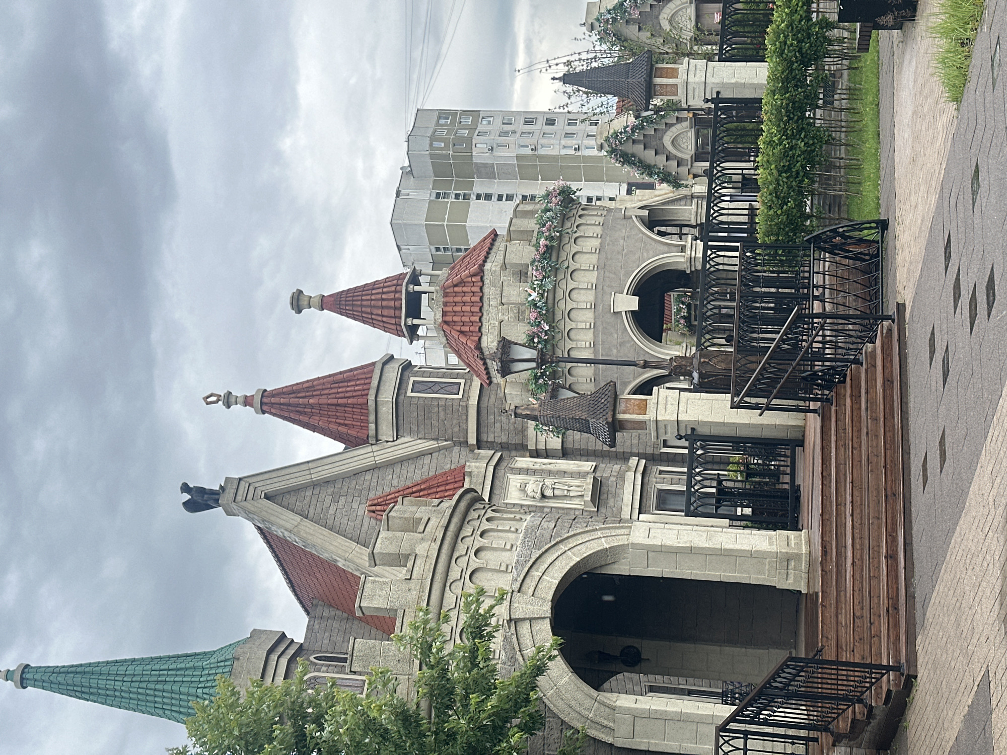 The image depicts a whimsical, castle-like structure with red-tiled conical roofs and grey stone accents. A bright green spire, reminiscent of a fairytale castle, rises prominently on the left. The building features arched doorways, decorative battlements, and what appears to be a statue of a knight in armor. Ornate black metal fences and railings, along with manicured green hedges, surround the structure. In the background, a tall, modern apartment building contrasts sharply with the fantastical architecture of the foreground. The sky is overcast, suggesting a cloudy day. This structure is the "Fantasyland" attraction located in the Ostankino Park in Moscow, Russia. No people are visible in the scene.