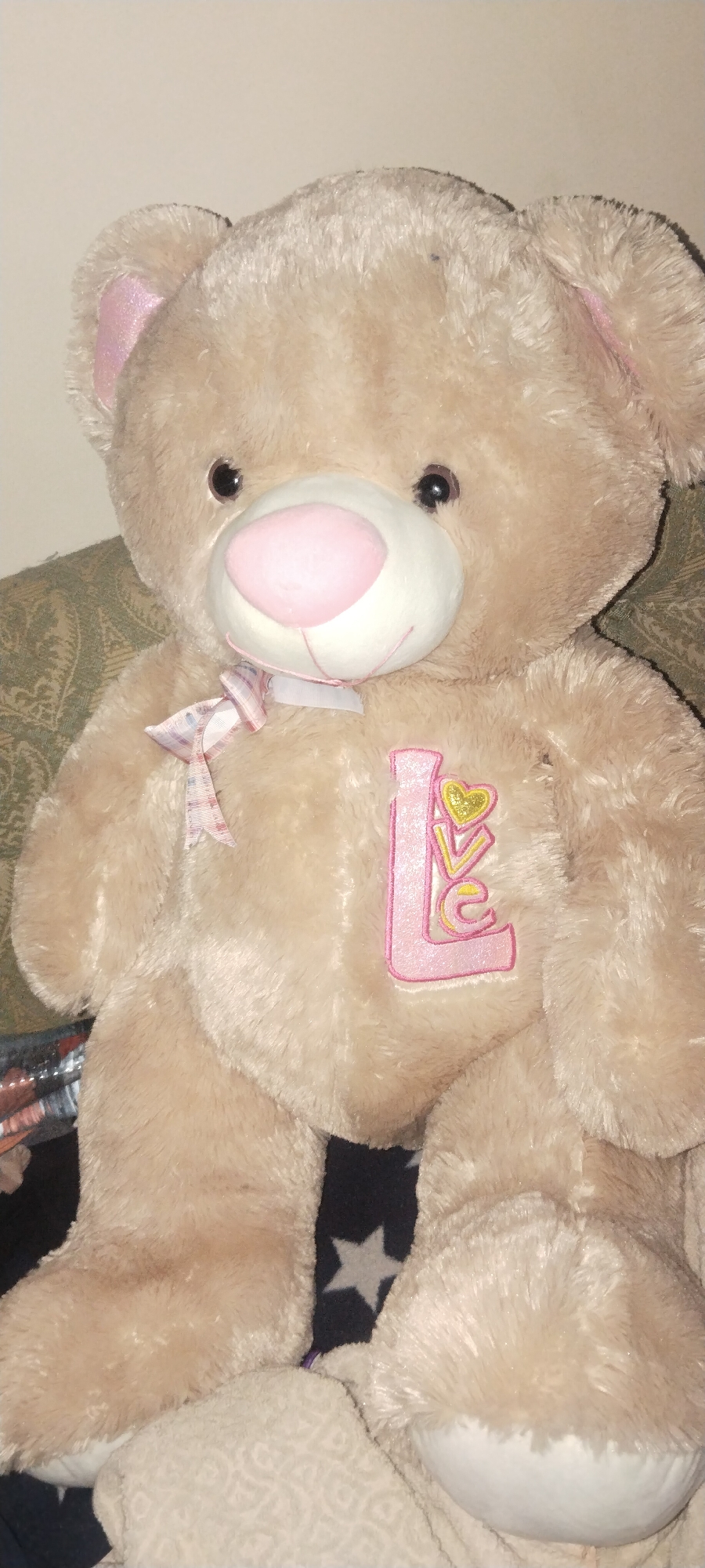 A tan teddy bear with pink ears and a pink nose sits on a couch. The bear has a pink ribbon tied in a bow around its neck. On the bear's chest is an embroidered patch that says "LOVE" in pink lettering, with a yellow heart. The bear is positioned in the center of the frame, filling most of the image. The background consists of a patterned green couch and a dark surface with stars on it. The lighting is dim, suggesting an indoor environment. There are no people visible in the image.