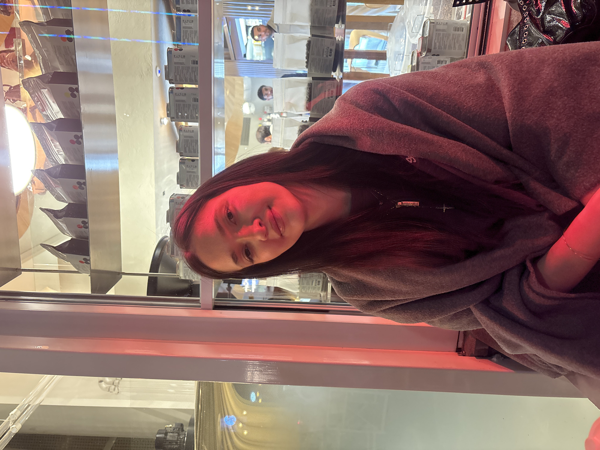 A woman is in Yerevan, Armenia, standing in front of a window displaying merchandise. The merchandise appears to be bags of coffee beans. The woman is wearing a gray shawl or cardigan. The lighting in the store appears to be red. Reflections of people and objects can be seen in the window.