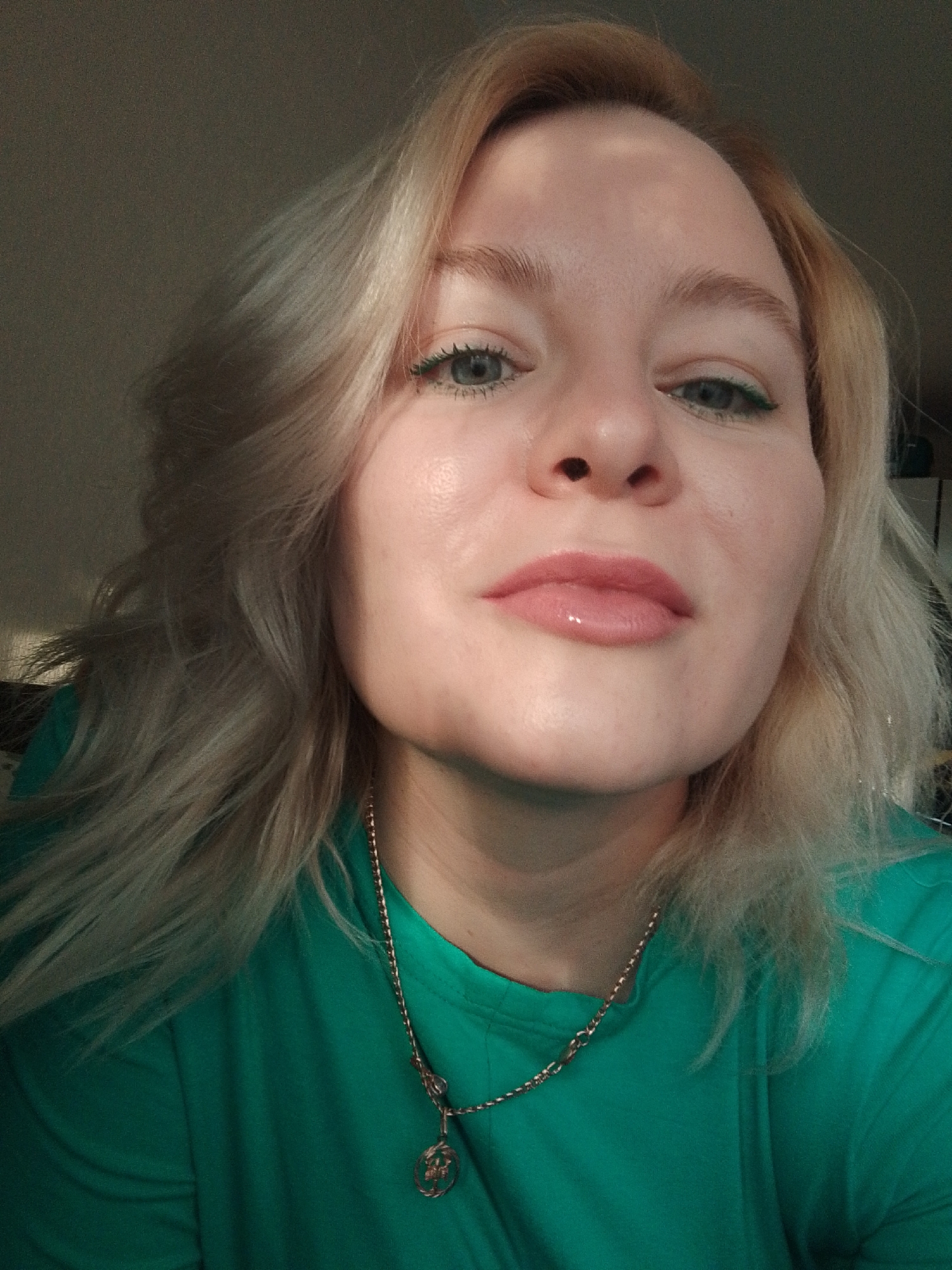 A close-up, high-angle shot features a woman with light blonde hair styled in waves. She is wearing a green t-shirt and a gold necklace with a circular pendant. Her makeup includes lip gloss and green eyeliner. The background is out of focus, but suggests an indoor setting with some light coming in from the left. There is no information to indicate that the location is in the city of Izhevsk, Russia.