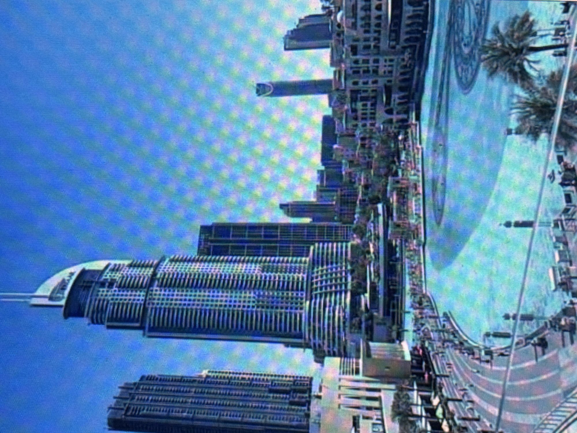 The image depicts a large, tiered skyscraper with a pointed top, situated in a city. To its left is another tall, rectangular skyscraper. In the foreground, a wide, circular body of water is visible, with a paved walkway surrounding it. Palm trees are scattered around the water's edge and on the walkway. The sky is a clear, bright blue. The scene appears to be a modern urban landscape with a prominent fountain or pool. There are no identifiable landmarks or features that would indicate the location is Pushkino, Russia.