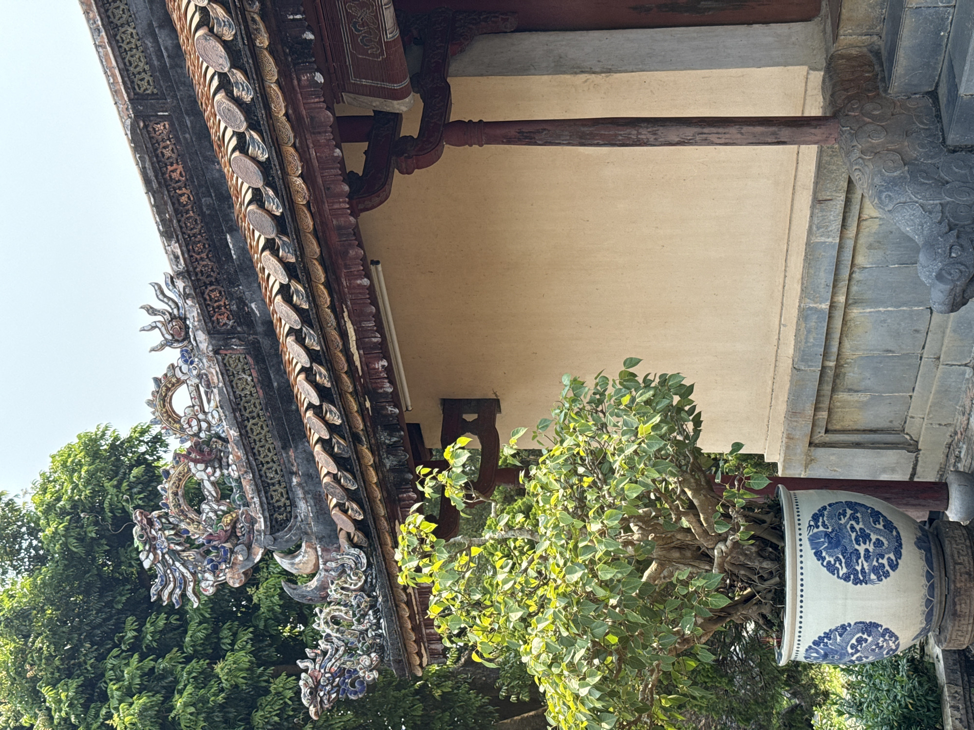 The image depicts a portion of a traditional Vietnamese building, likely a temple or palace structure, situated in Kim Long Ward, Vietnam. A section of the building's ornate roof is prominently displayed, featuring intricate carvings, decorative tiles, and a multi-tiered design. The roof is adorned with representations of mythical creatures, possibly dragons, and detailed geometric patterns.
Below the roof, a section of the building's wall is visible, painted a light beige color. Wooden support beams and brackets are used to brace the roof structure. A large ceramic pot, decorated with blue floral and possibly dragon motifs, sits in the foreground. A leafy green tree grows out of the pot, partially obscuring the view of the building.
The image suggests an architectural style specific to Vietnamese cultural heritage, with a focus on ornate details and traditional building materials. The presence of the bonsai tree suggests that these buildings are also decorated with plants.