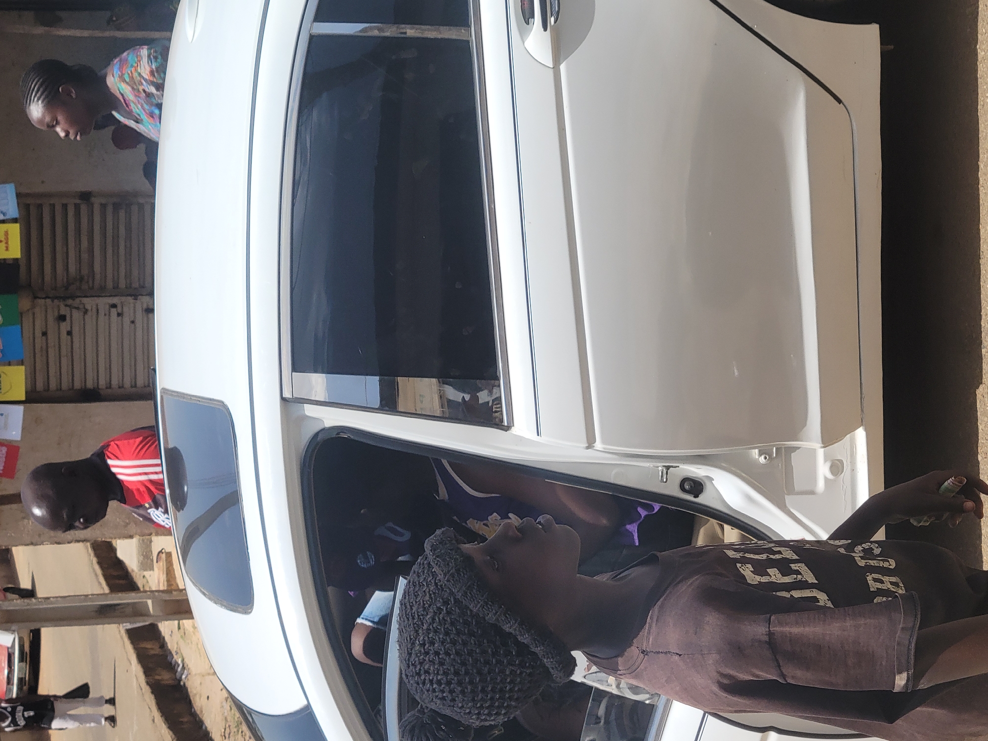 The image shows a white car with its door open on a street in Jos, Nigeria. Several people are visible around the car. One person is looking into the car while wearing a purple shirt. Another person stands outside the car, dressed in a brown shirt and knitted cap, holding something in their hand. Additional individuals are partially visible, including someone in a red shirt looking into the rear window of the car, and another person is looking into the front window. Background includes a building with signs, suggesting a commercial area.