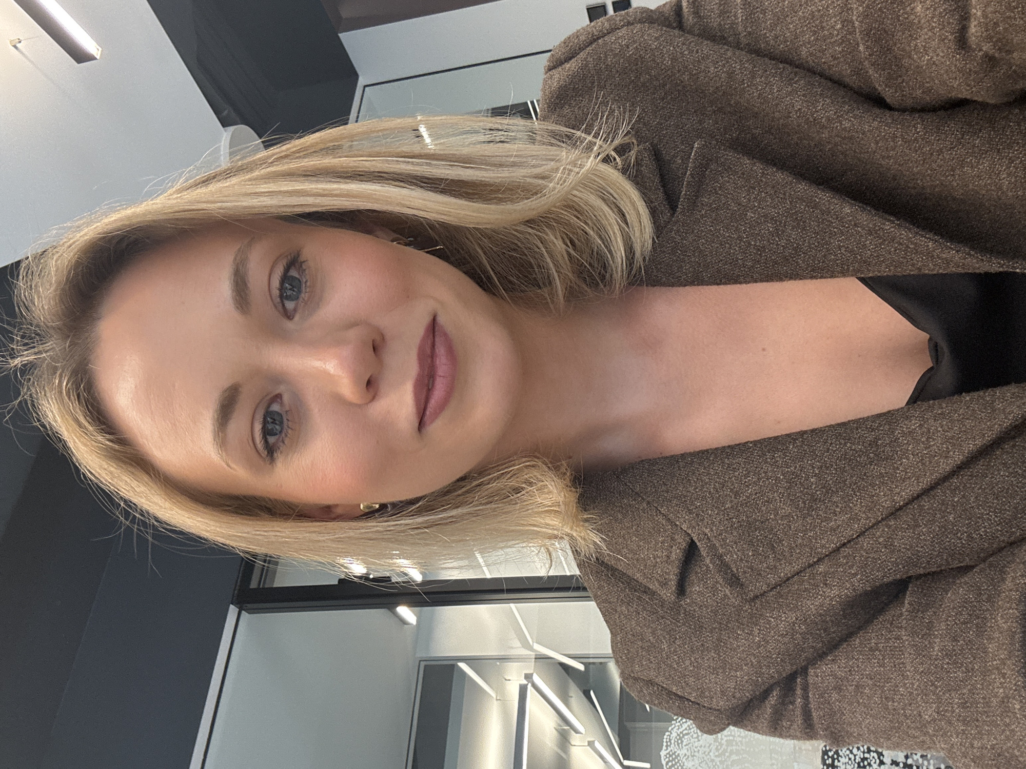 A medium close-up, portrait style photograph shows a blonde-haired woman wearing a brown tweed blazer. The blazer is textured and buttoned. Underneath the blazer, a black garment is visible. The woman has fair skin, light eyes, and is wearing makeup on her lips and cheeks. She is gazing directly at the camera with a slight smile. The background is blurred and appears to show an office or commercial space with neutral grey, white, and black colors. There are geometric lighting fixtures present in the background. The location details (Riga, Latvia) cannot be definitively determined based solely on the visual information in the image.