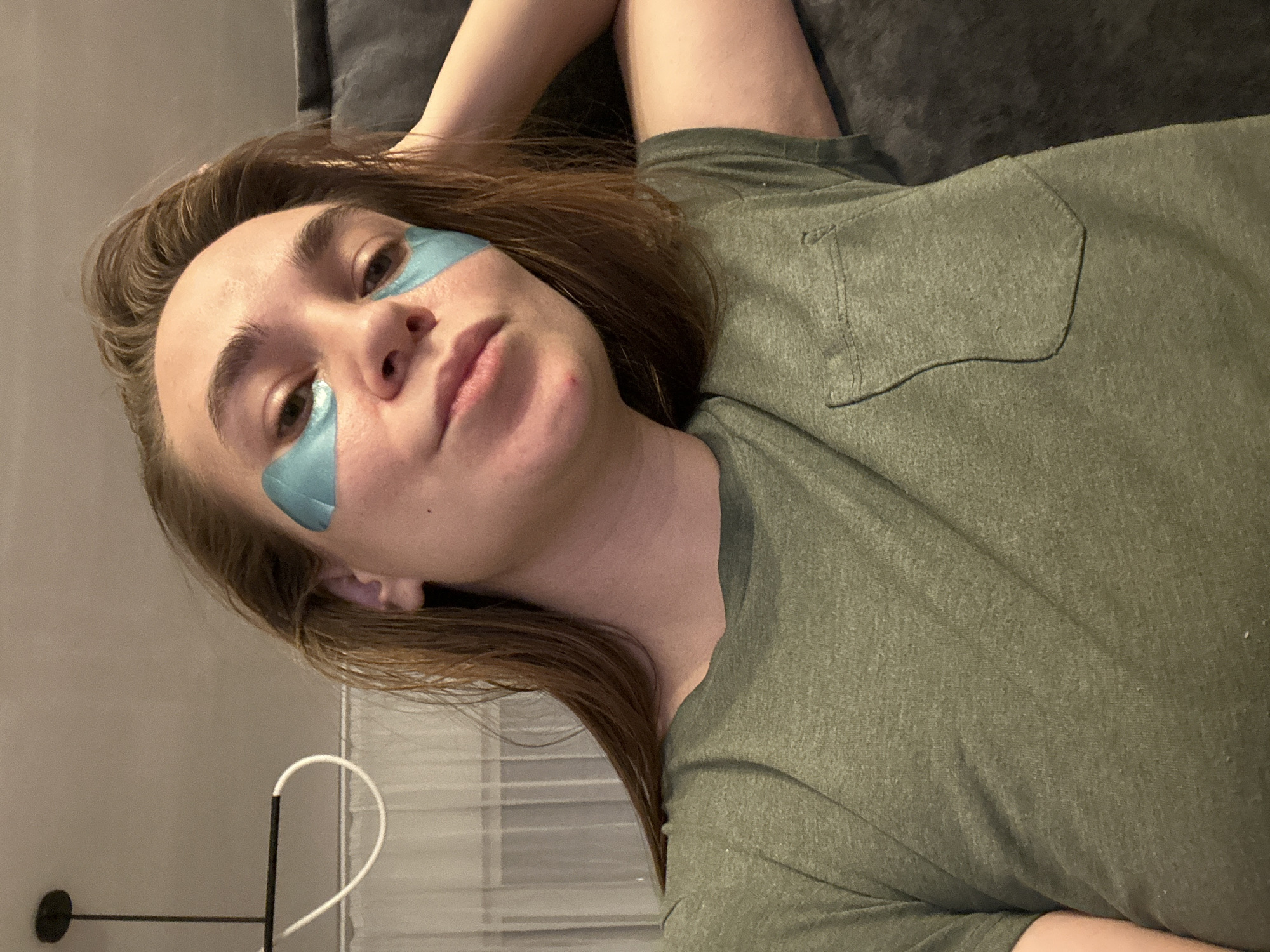A woman lies on a gray surface, likely a couch or bed, wearing blue under-eye patches. She is wearing a green t-shirt with a pocket. Her hair is brown. In the background, there is a white cord and what appears to be blinds. There is no evidence indicating the location of Kharnavy, Russia.