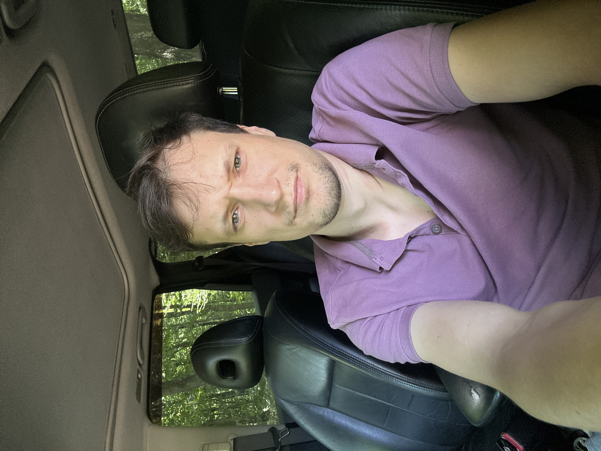 A man with short brown hair and light facial hair is taking a selfie inside a car. He is wearing a purple polo shirt. The car has black leather seats and a sunroof. Through the windows, trees and foliage are visible, suggesting an outdoor, possibly rural, location. The specific location of Shatava, Ukraine, is not determinable from the image itself.
