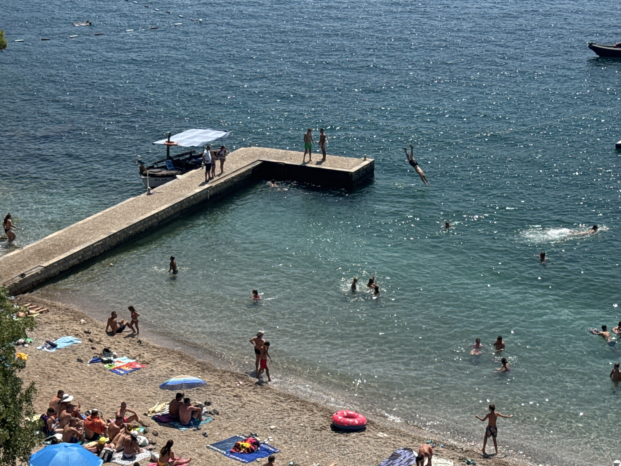 This is an aerial photograph of a beach and its adjacent sea, located in Budva, Montenegro. The scene depicts a sunny day with a clear blue sky and calm, shimmering water.
On the sandy beach, numerous people are sunbathing and relaxing on towels and under umbrellas. Some are seated, while others are lying down. A group of people are visible near the water's edge, some wading in the shallow sea. Beach accessories such as towels, bags, and umbrellas are scattered across the sand.
Extending from the beach into the water is a concrete pier. On the pier, several individuals are standing, and one person is captured mid-air, diving into the sea. A small boat with a canopy is docked at the pier.
The sea is populated with swimmers. Several people are floating or swimming in the clear turquoise water. A few more are further out, and one person is visible near a dark boat in the distance. The water is shallow near the shore, gradually deepening as it extends out. The overall atmosphere is one of leisure and recreation on a summer day.