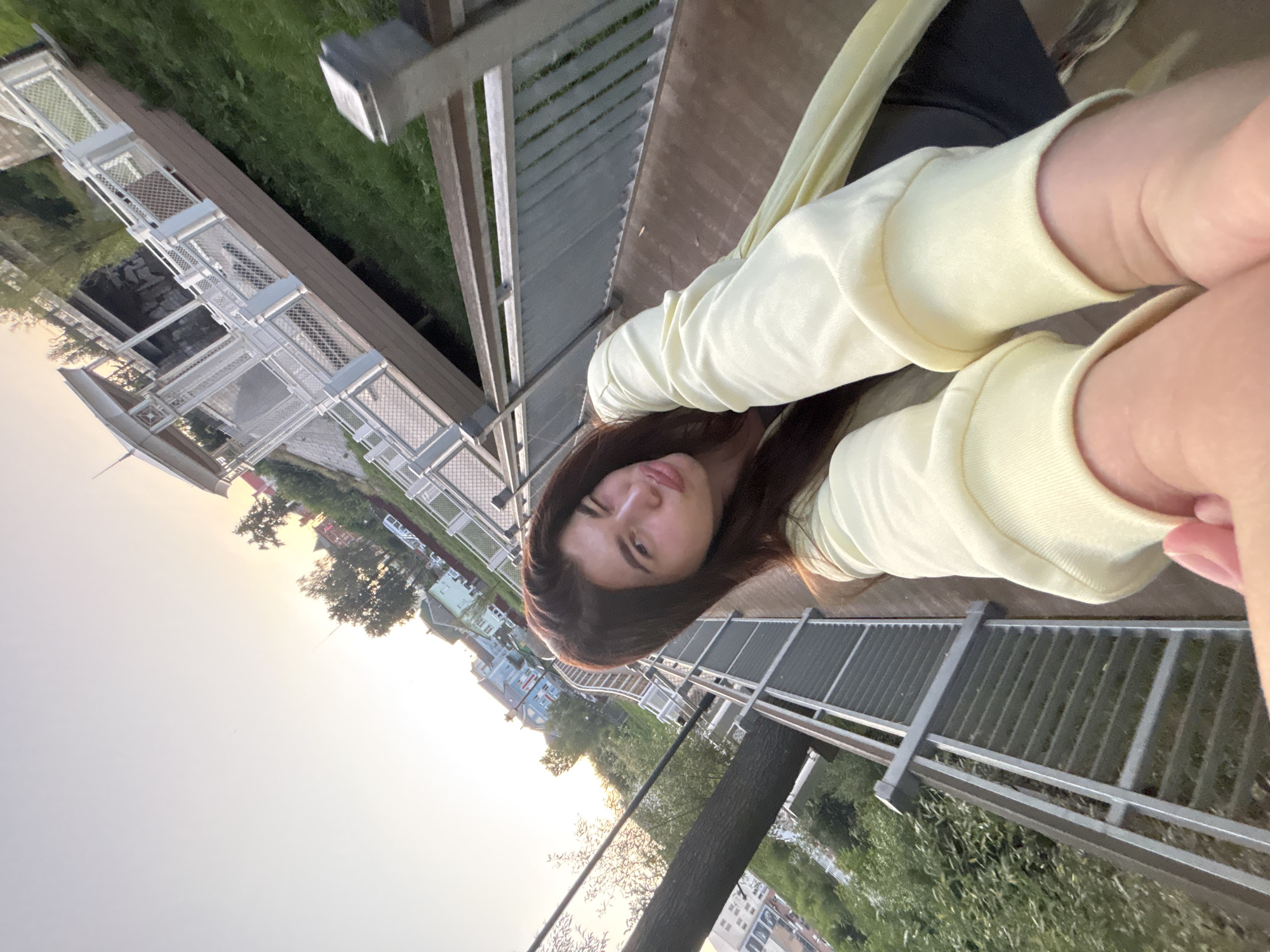 A young woman with long brown hair is taking a selfie on a boardwalk in Klin, Russia. She is wearing a yellow sweatshirt and has one eye closed in a wink. The boardwalk has metal railings and wooden planks. In the background, there are trees, houses, and a gazebo. The sun is setting, casting a warm glow over the scene. The image is taken from a low angle, with the woman's arm extending towards the camera.
