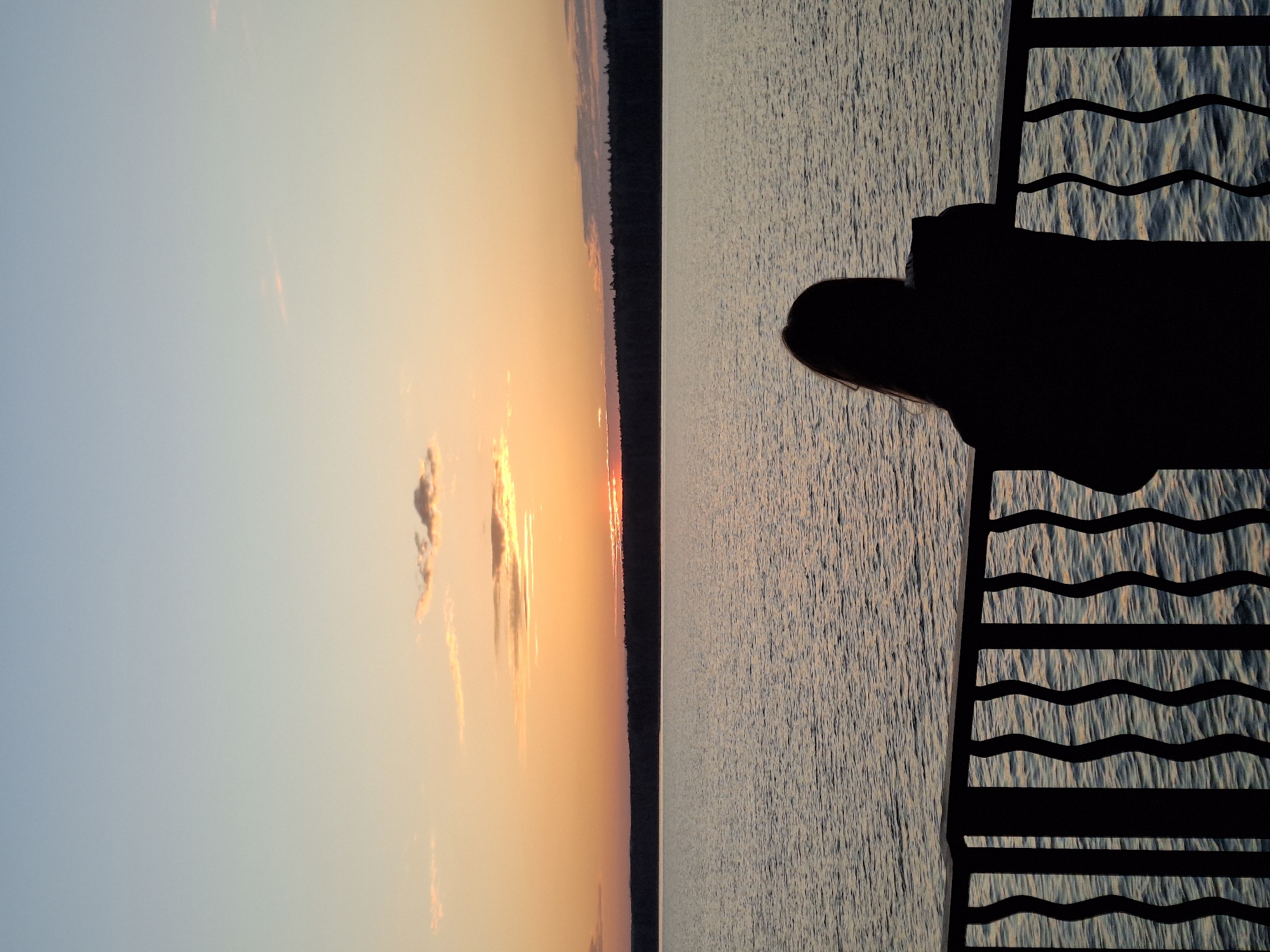 A silhouette of a person is seen sitting in a chair facing the lake, the setting sun and the distant tree line at Chaikovsky, Russia. The colors are soft, with warm hues in the sky. The water surface is textured by ripples, reflecting the sky. A metal railing is visible on the right.
