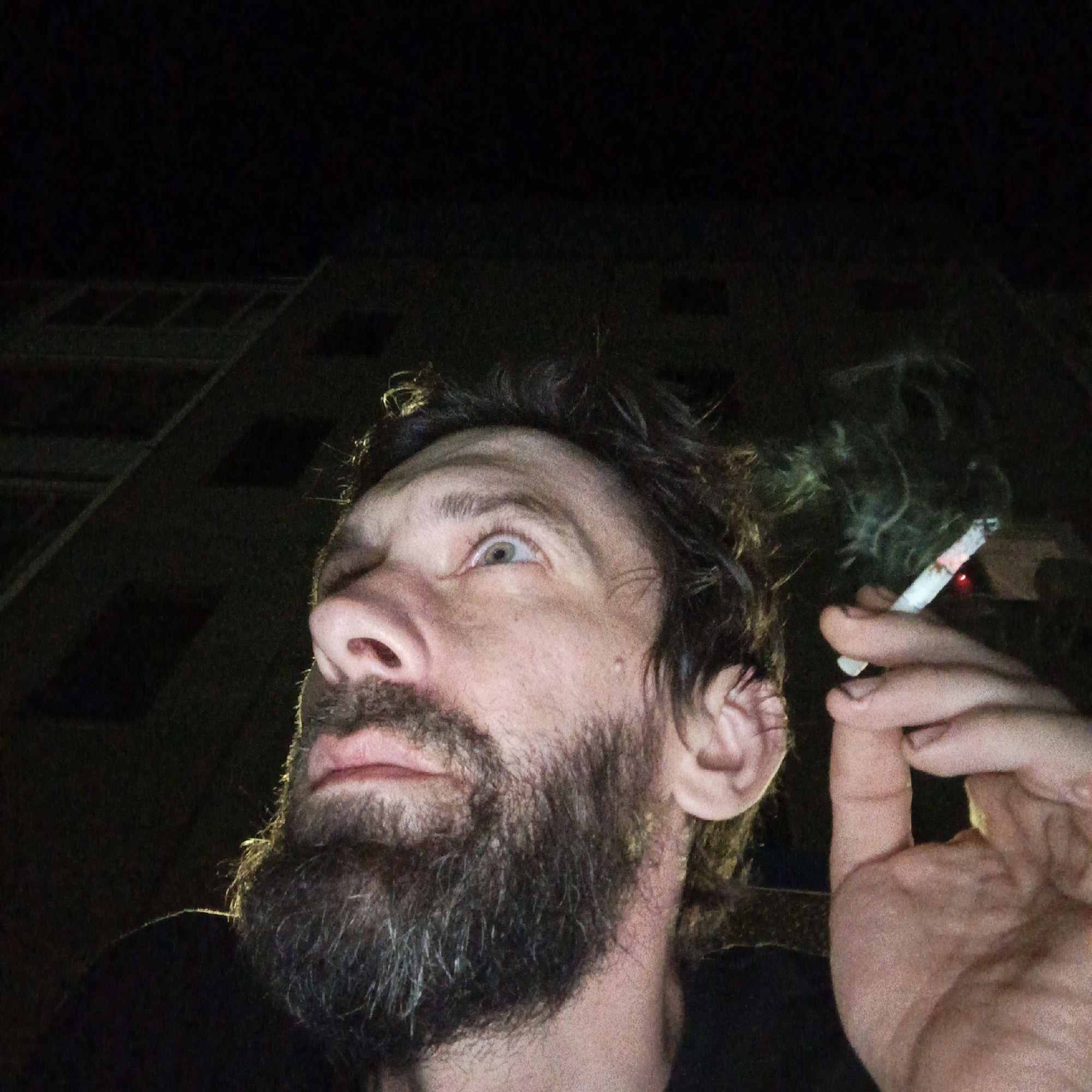 A bearded man is holding a lit cigarette. He is looking upwards. It is nighttime. A building with lighted windows can be seen behind the man. There is smoke emanating from the cigarette. It is not possible to verify the location in the city of Марковский, Russia from the available visual information.