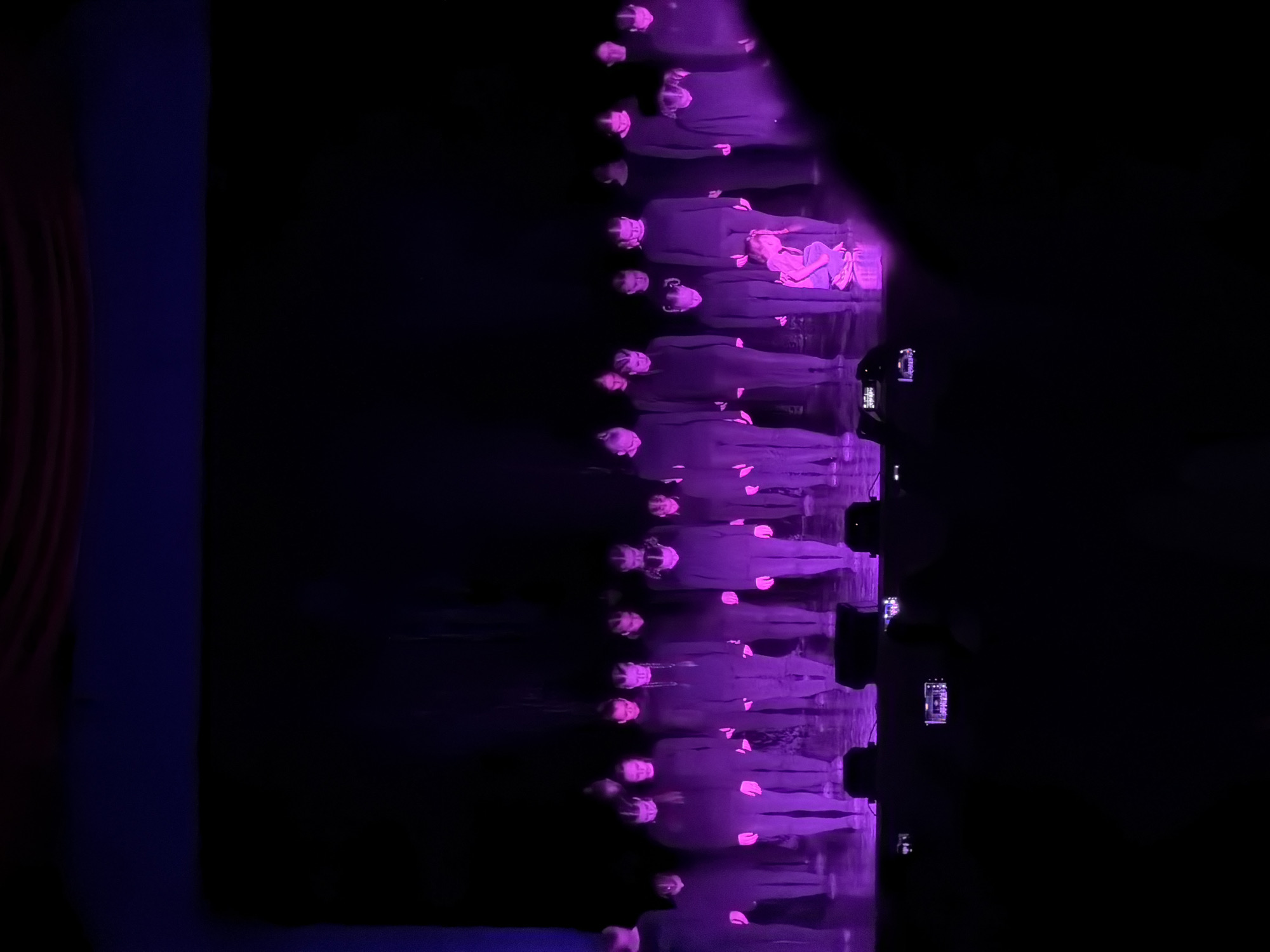 The image captures a performance or event in a dimly lit venue, likely a theater or concert hall, with strong purple and magenta stage lighting illuminating a group of people. The composition is predominantly dark, with the light focused on the right side of the frame.
The main subjects are numerous individuals, primarily adults, arranged in multiple horizontal rows on what appears to be a stage or elevated platform. They are dressed in dark, possibly uniform-like attire. Their faces are illuminated by the vibrant purple light, and their expressions suggest concentration or involvement in a performance. Among the adults, a child is visible towards the front-right of the illuminated group, wearing a lighter-colored garment and appearing to be lying down or held.
The activity taking place is a staged performance, possibly by a choir or an ensemble, given the structured arrangement of the participants. On the far right, beyond the main illuminated area, several figures in the background are visible, holding up what appear to be smartphones or cameras, indicating an audience recording the event.
The notable details include the dramatic and colorful lighting, which creates a striking visual effect against the dark background. The scene suggests an evening event indoors. No discernible text is visible within the image. The location context provided for this event is Perm, Russia.