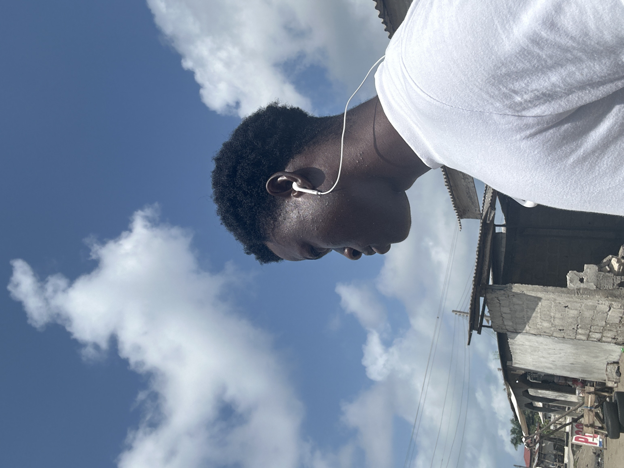 The image, rotated 90 degrees clockwise, captures a close-up profile of a young person, likely male, with dark skin and short, curly black hair. He is wearing a white t-shirt and white wired earphones are visible in his right ear, with the wire extending downwards. His face is turned downwards and to the right, with prominent cheekbones and what appears to be sweat or skin texture visible on his face and forehead.
The background is dominated by a bright blue sky with scattered white cumulus clouds. In the upper and lower right portions of the frame, behind the person, an urban or semi-urban environment is visible. Structures made of concrete blocks, some appearing weathered or under construction, are present, topped with what looks like corrugated metal roofing. Utility wires are strung across the mid-ground. At the very bottom right, a small red and white sign with the partial text "PE" can be seen.
The scene depicts the person outdoors on a bright, sunny day, suggested by the strong light and clear sky. The setting, with its building styles and utility lines, aligns with the provided location context of Badagry, Nigeria. The activity appears to be listening to music or audio through earphones while in this outdoor setting.