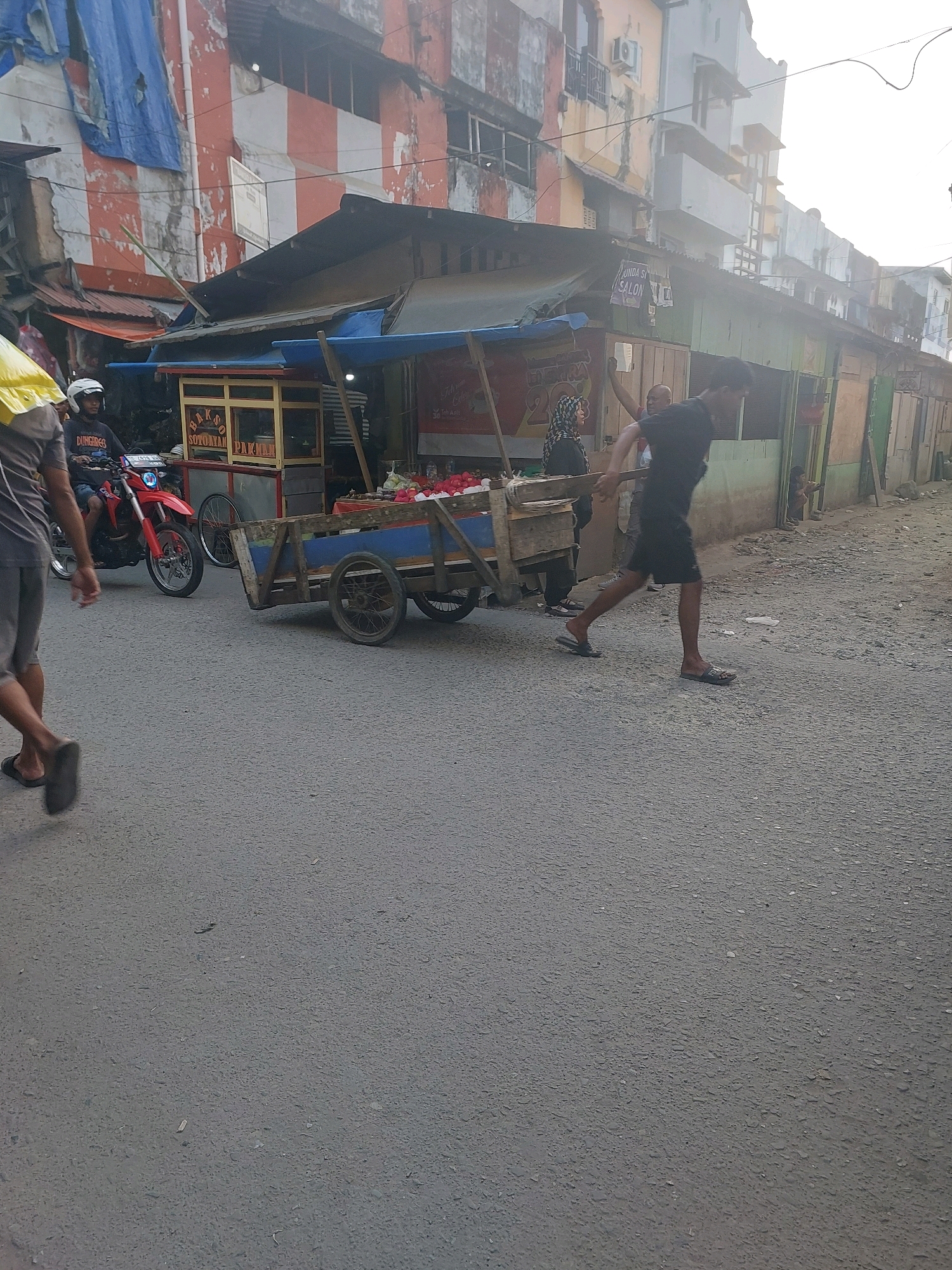 This image captures a bustling street scene in Ambon, Indonesia, likely in a market area or a residential neighborhood with commercial activity. A red motorcycle with a rider wearing a white helmet and a dark shirt is parked on the left side of the road. Next to it, a food cart selling "BAKSO SOTOYAN PAKMAN" is visible, with red and yellow branding. A man in a black t-shirt and shorts is pushing a wooden cart filled with white and red items, possibly flowers or produce, towards the right side of the frame. His movement suggests he is engaged in an activity related to the cart's contents or transport.
In the background, several buildings line the street. The closest building on the left has a weathered facade with red and white stripes and blue tarps draped over its upper section, indicating ongoing construction or disrepair. Further down the street, a row of buildings with various colored walls, including green and beige, are visible. Some have awnings or open fronts, suggesting shops or stalls. The street itself is paved with asphalt and appears to be a public thoroughfare. The overall atmosphere is one of daily life and commerce in an urban Indonesian setting.