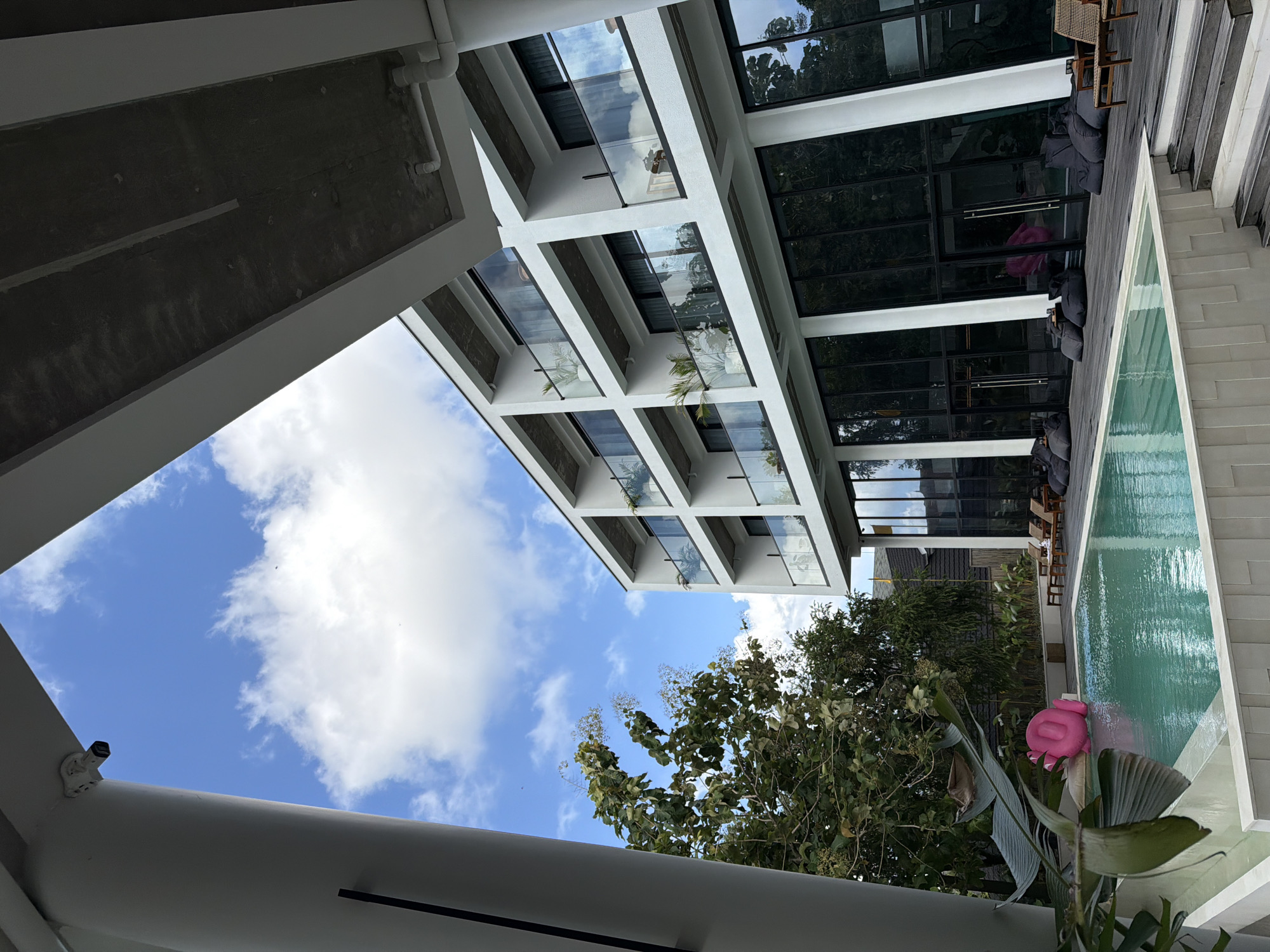The image shows an upward view of a modern building complex in Mengwi, Indonesia. The building has a white facade with multiple levels of balconies featuring glass railings. Sections of the building have glass roofs supported by white columns, providing partial shelter over a swimming pool area. The pool is surrounded by wooden decking and lounge furniture, including chairs and beanbag-style seating. A large tropical plant with broad leaves and a pink inflatable object is visible near the pool. The sky is blue with scattered white clouds, visible through openings in the building structure. Trees and other vegetation are visible in the background, suggesting a tropical environment.